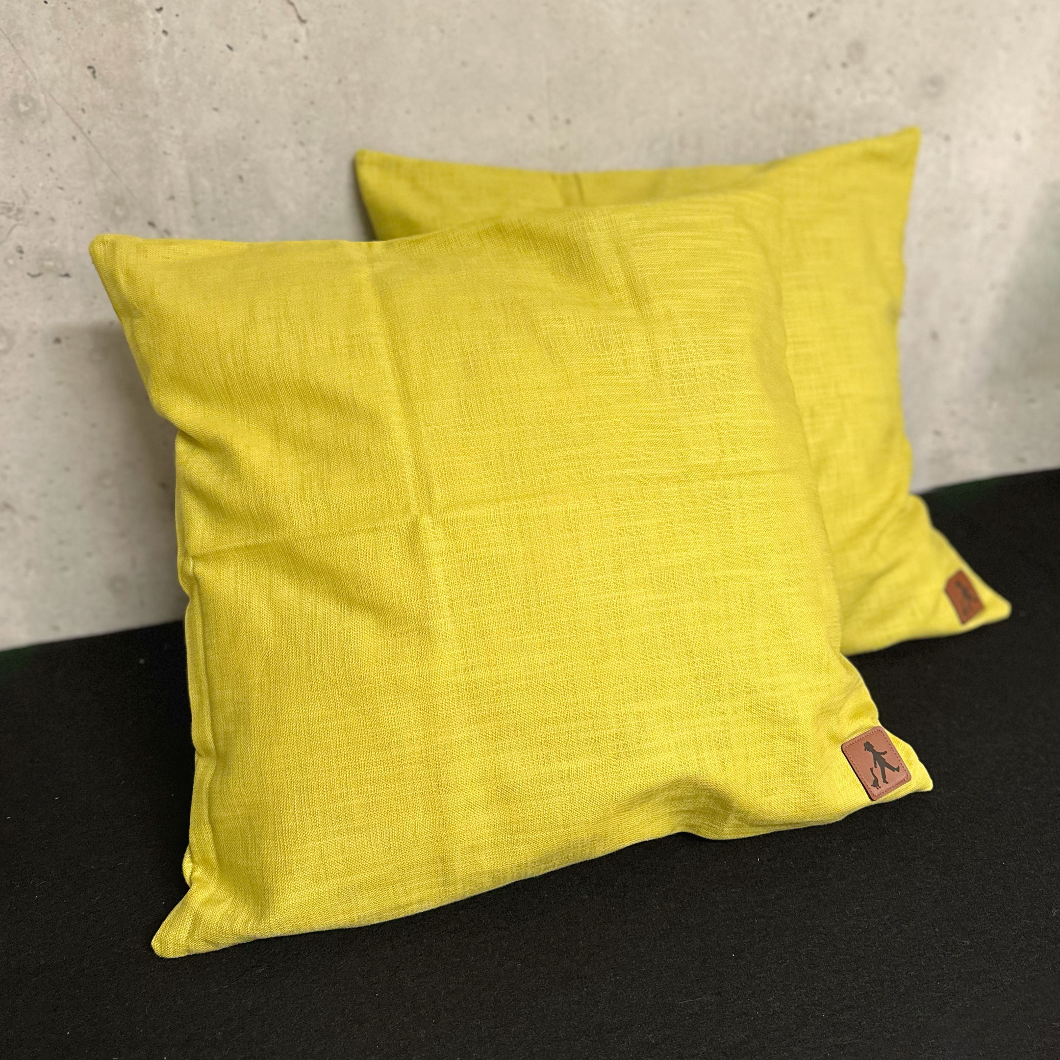 Zwei gelbe Kissen liegen auf einem dunklen Sofa mit Betonwand im Hintergrund – betont den wohnlichen Charakter und die Stoffstruktur.
