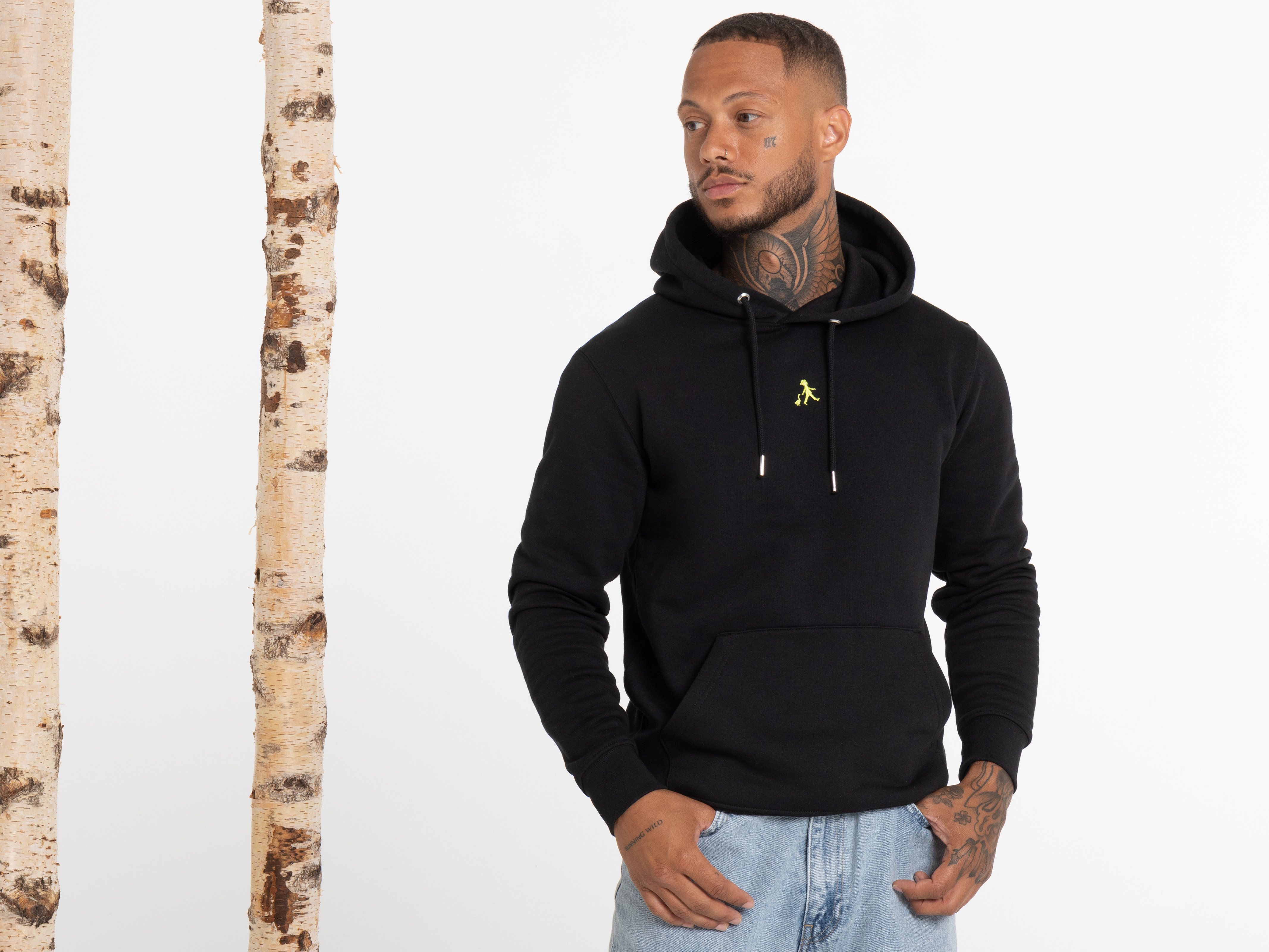 Ein Mann steht frontal und trägt einen schwarzen Hoodie mit geschlossenem Reißverschluss und Kapuze. Er hat die rechte Hand in der Hosentasche und blickt leicht zur linken Seite. Die Pose wirkt selbstbewusst und ruhig.