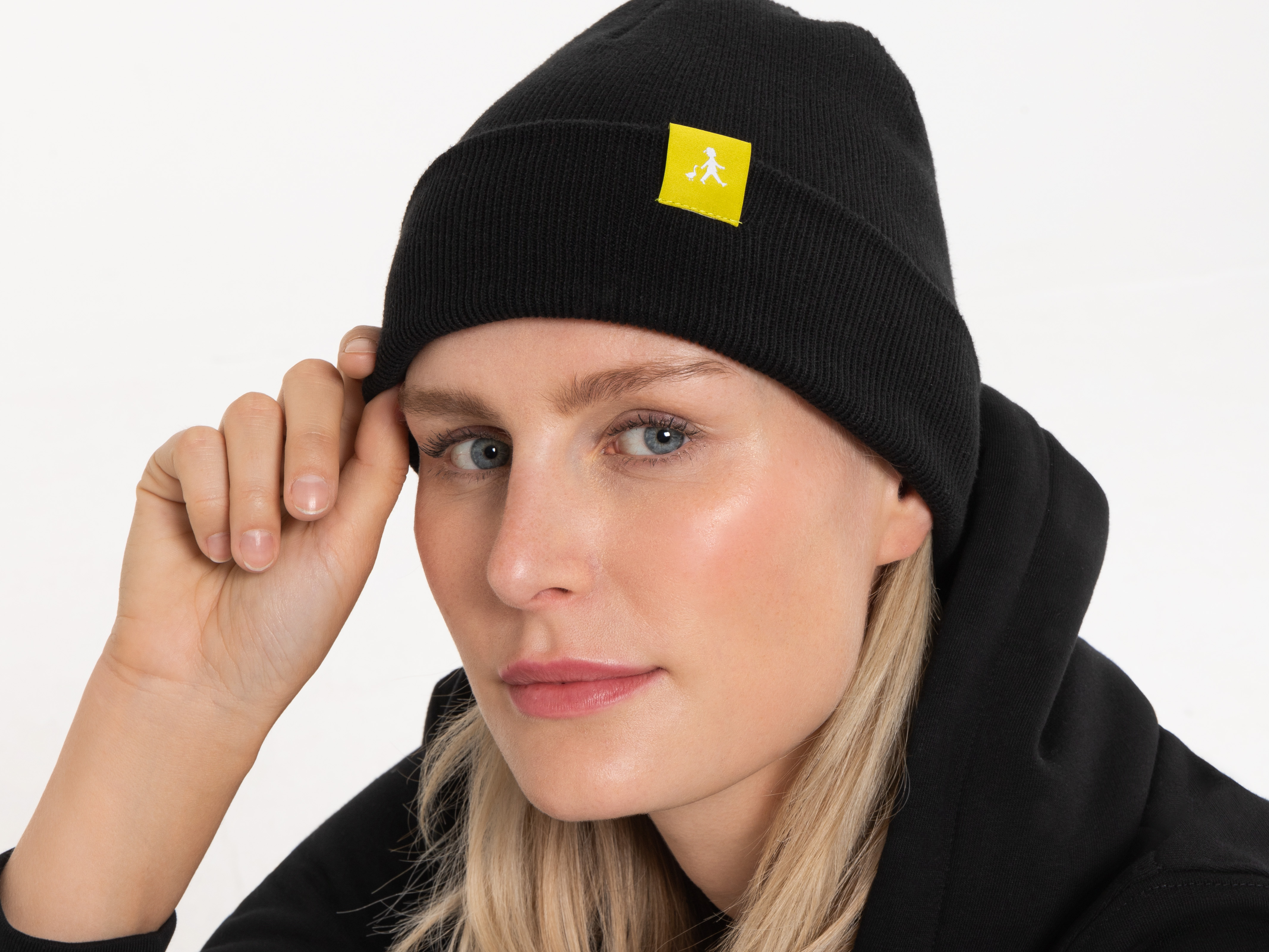 Porträtaufnahme einer Frau mit schwarzer Beanie mit gelbem Logo. Sie schaut zur Seite und hält die Kappe leicht mit einer Hand fest.