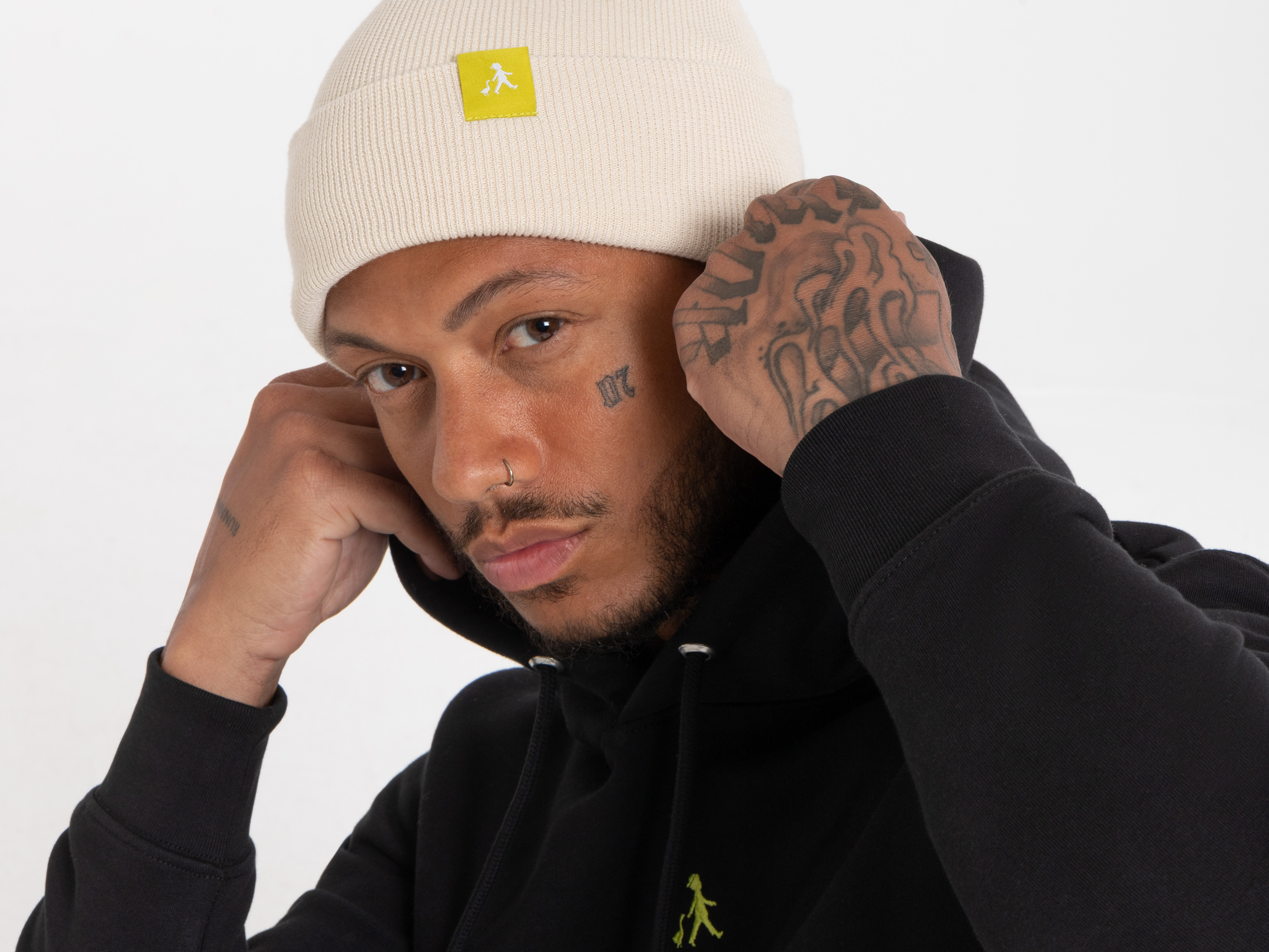Frontalaufnahme eines Mannes mit hellbeiger Beanie und gelbem Label. Er trägt schwarzen Hoodie und blickt ernst in die Kamera.