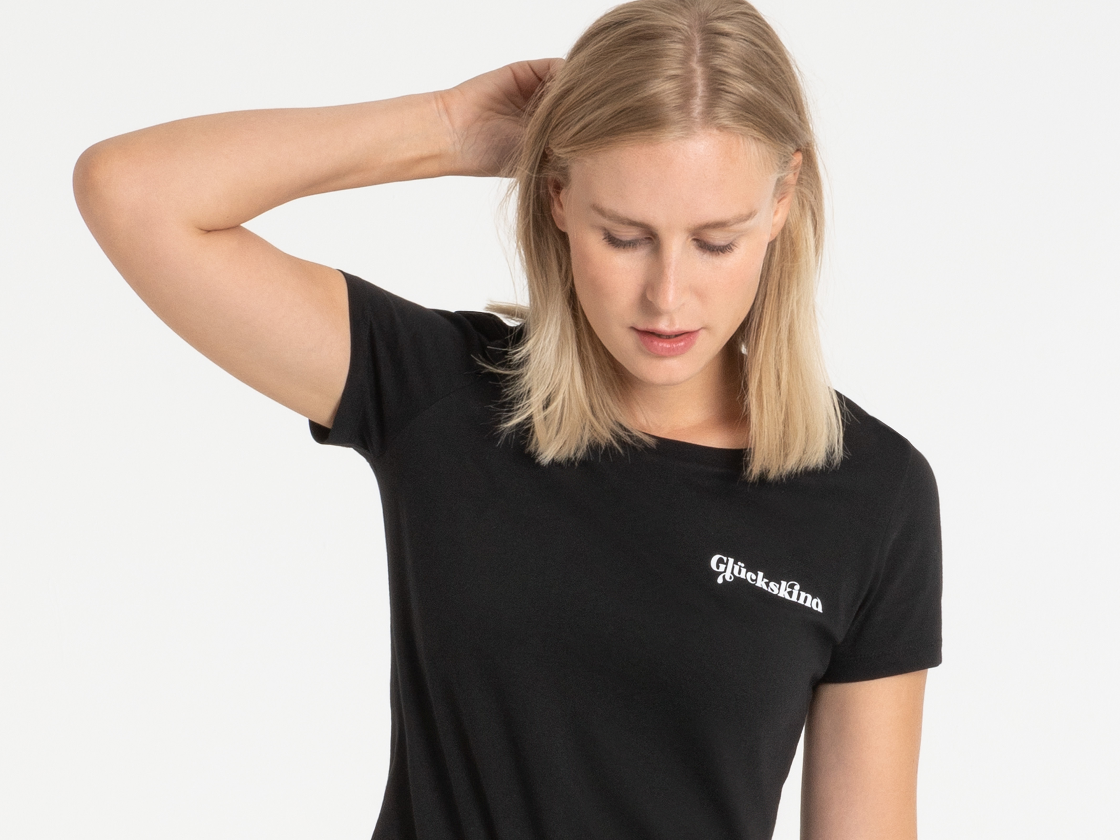 Frau mit lockerem Griff ans Haar, trägt schwarzes T-Shirt mit kleinem Frontlogo – natürlicher Streetstyle.