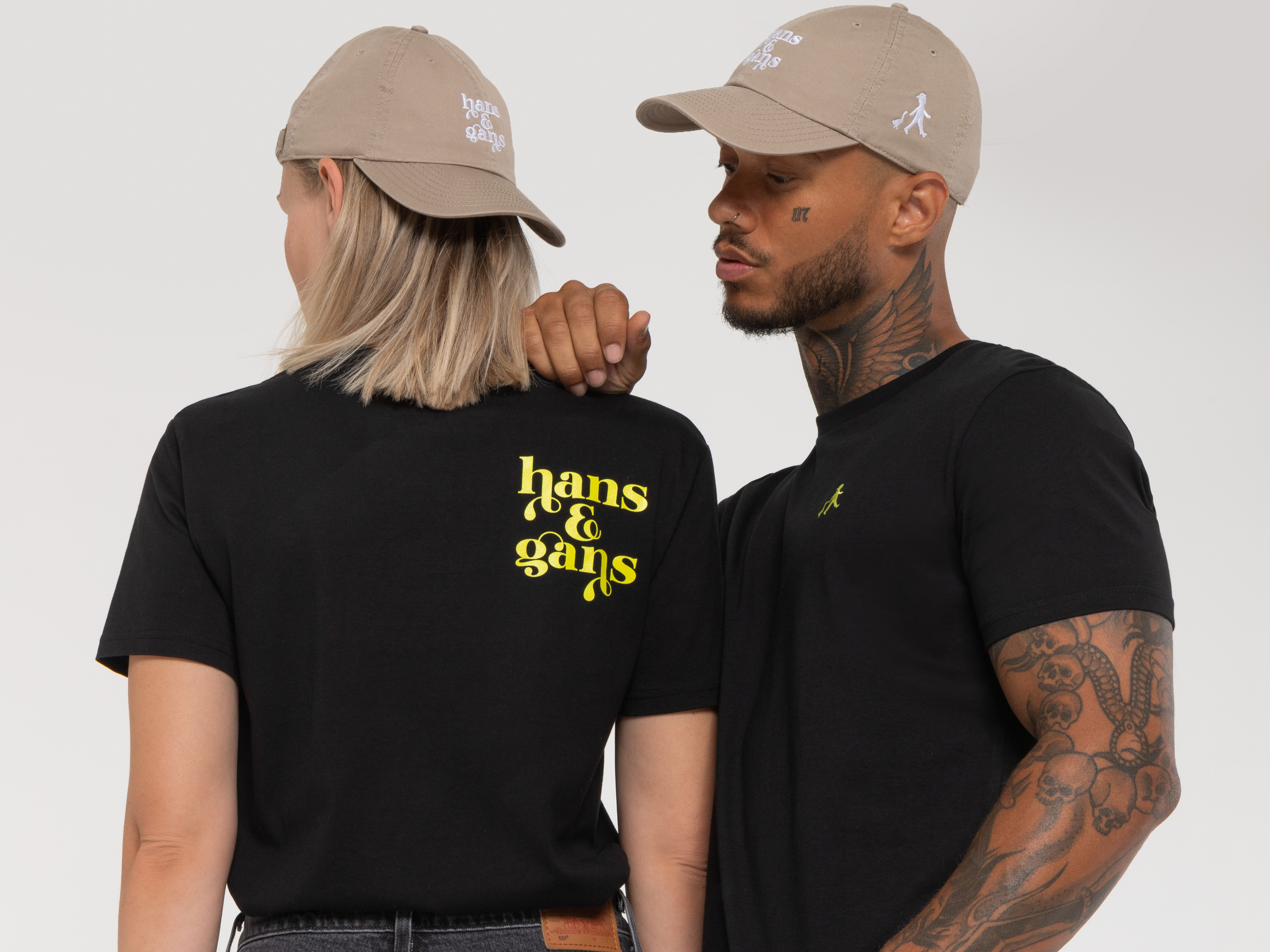 Zwei Personen stehen Rücken an Rücken. Beide tragen schwarze Shirts, die Frau eine beigefarbene Cap und der Mann ein schwarzes Shirt mit gelbem „hans & gans“-Print.