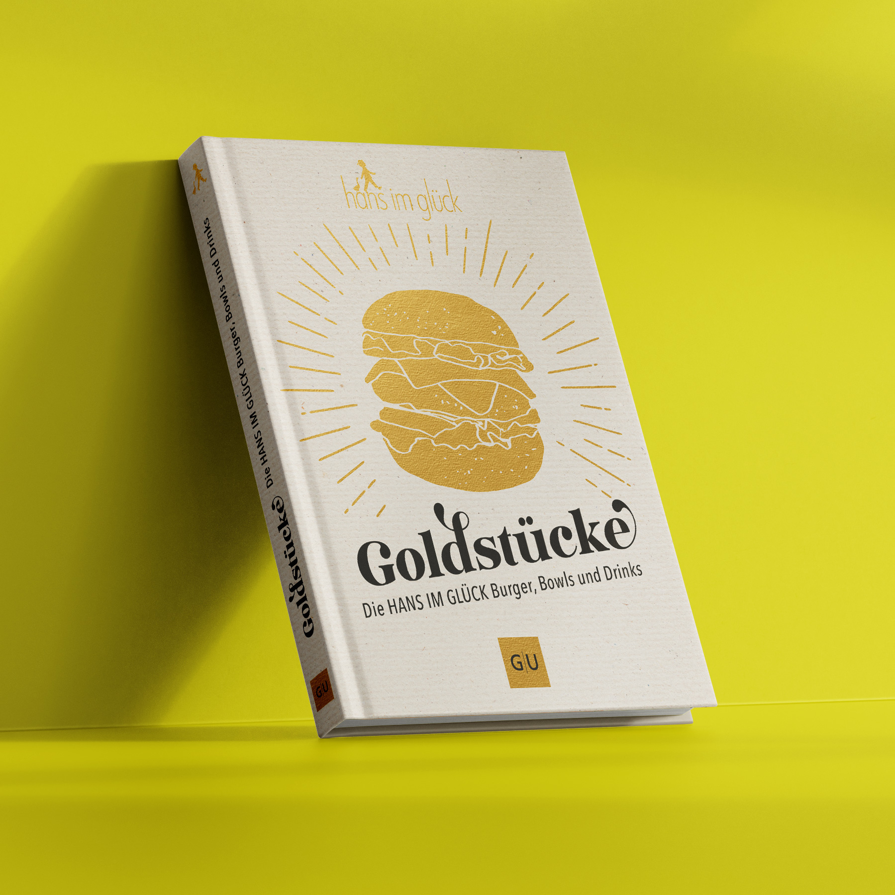 Geschlossenes Kochbuch „Goldstücke – Die HANS IM GLÜCK Burger, Bowls und Drinks“ auf gelbem Hintergrund, cremefarbener Leineneinband mit goldenem Burger-Illustrationsmotiv, Verlagslogo „GU“ am unteren Rand, klare Produktaufnahme des Buchcovers.