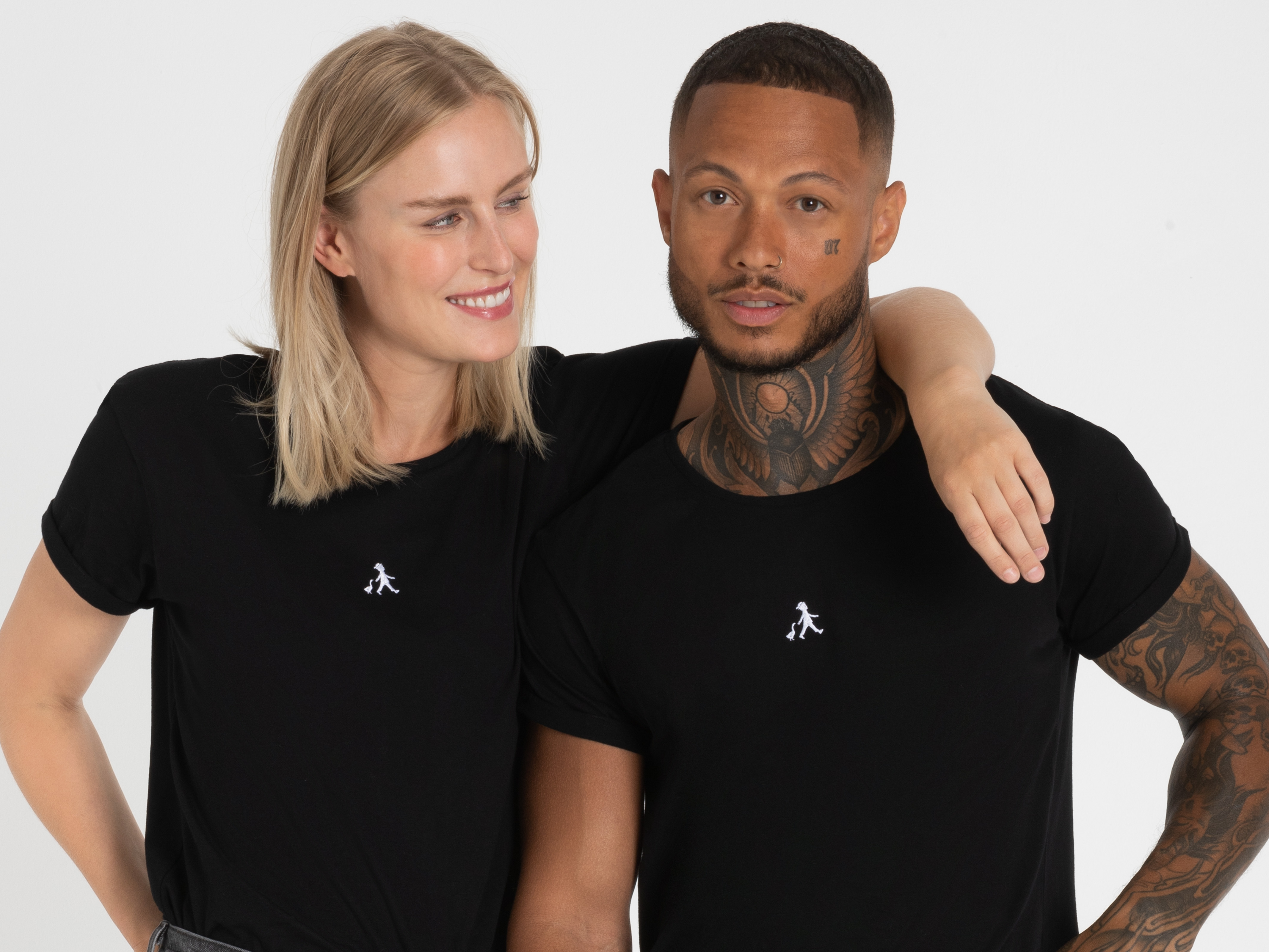 Zwei Personen, eine blonde Frau und ein dunkelhäutiger Mann, stehen Schulter an Schulter in schwarzen Poloshirts und lachen herzlich.