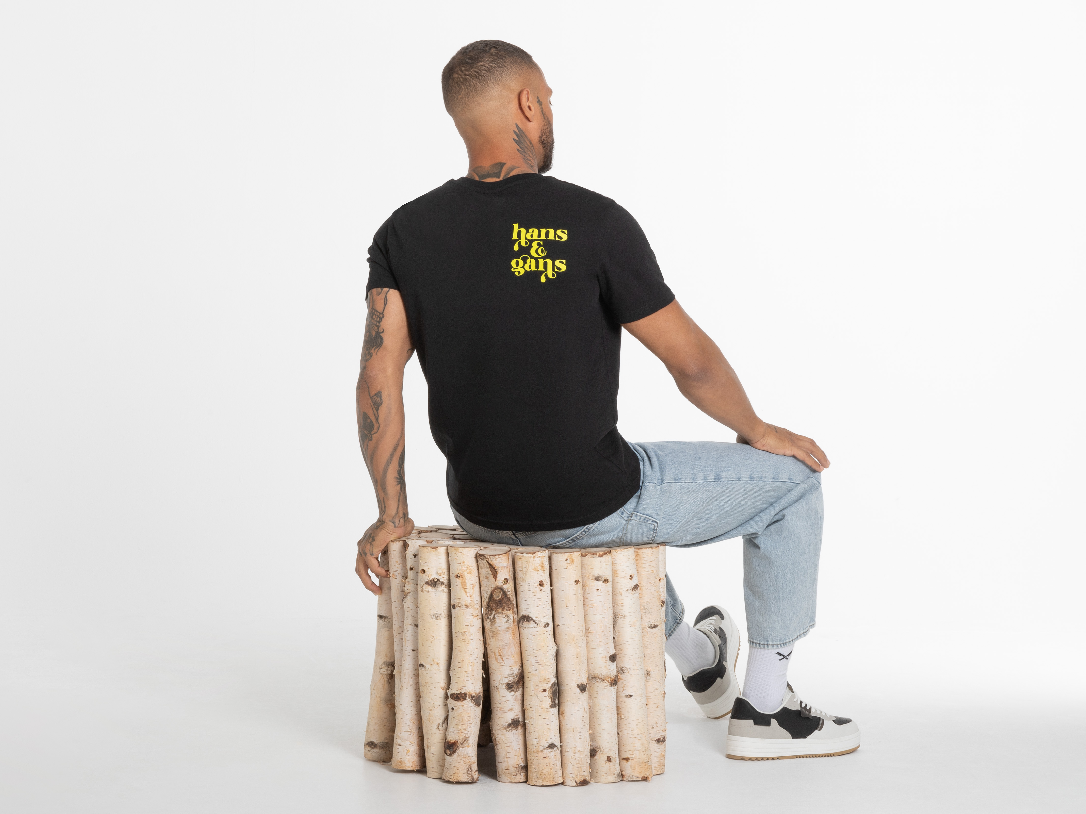 Mann sitzt seitlich auf Holzstamm, zeigt Rückenprint des schwarzen Shirts – cooles Outfit für lässige Momente.