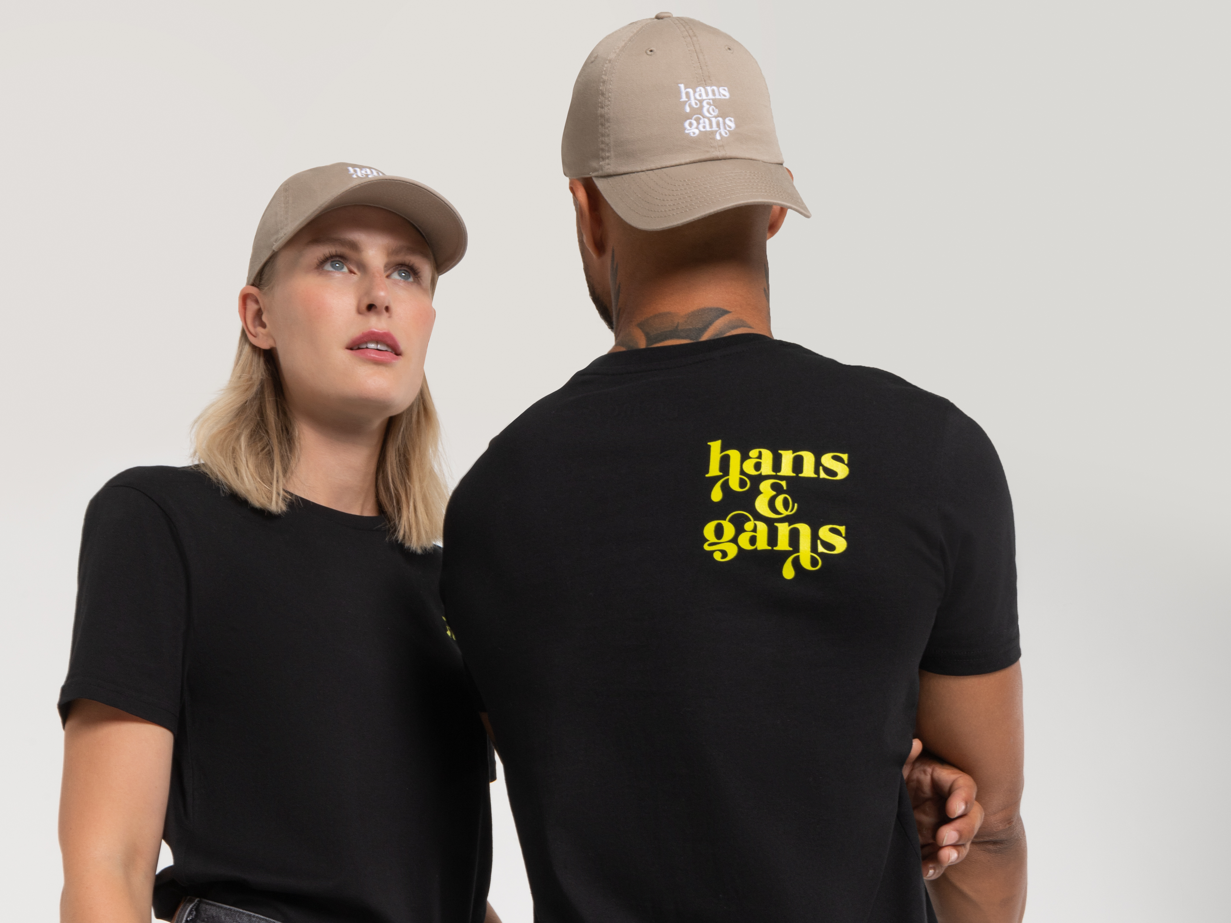 Zwei Personen stehen Rücken an Rücken. Der Mann trägt ein schwarzes T-Shirt mit gelbem Rückenprint „hans & gans“, die Frau eine beigefarbene Cap – modische Kombi mit Markenbezug.
