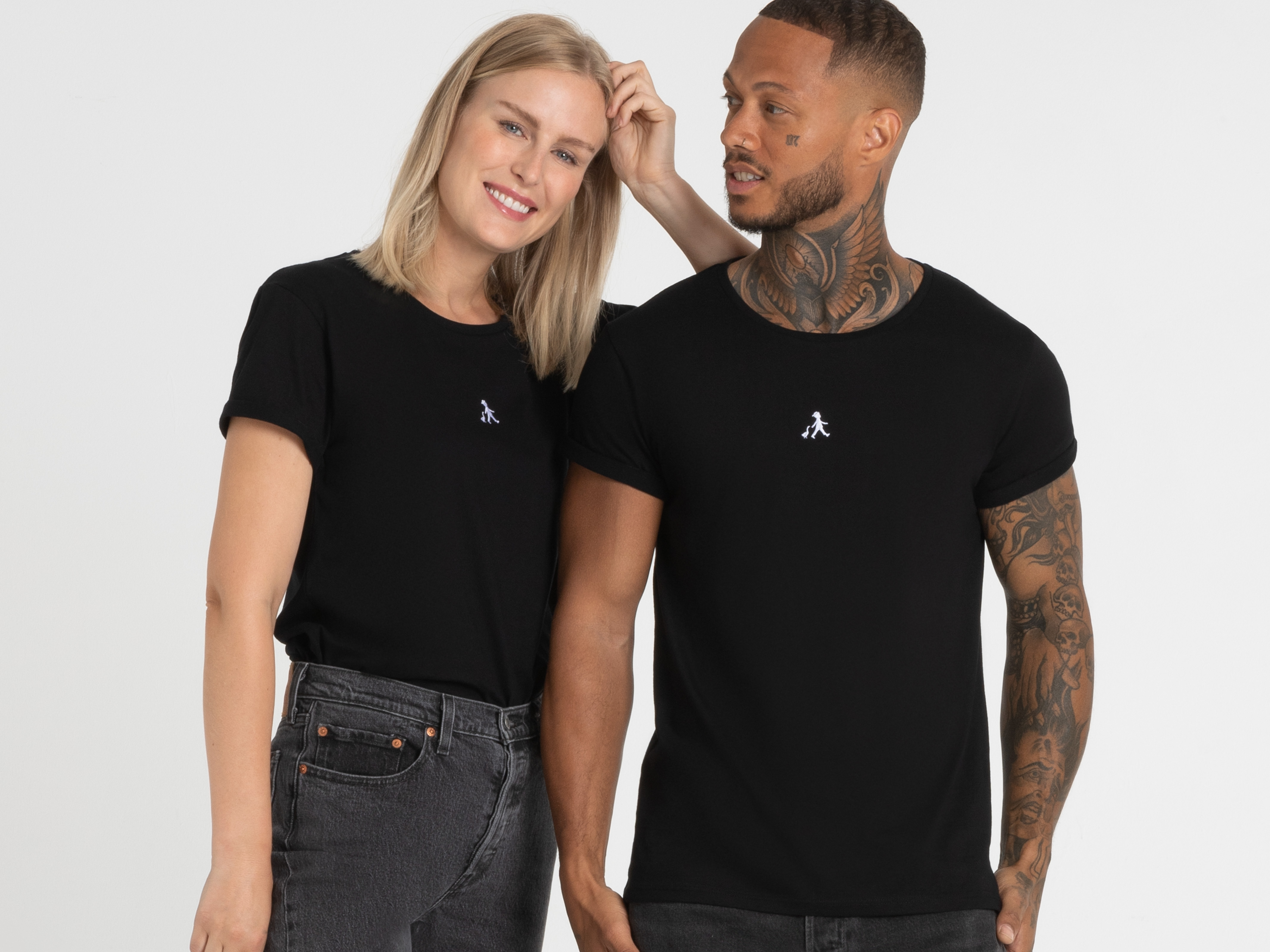 Eine blonde Frau mit Mittelscheitel steht entspannt neben einem dunkelhäutigen Mann, beide tragen schwarze Shirts und blicken selbstbewusst in die Kamera.