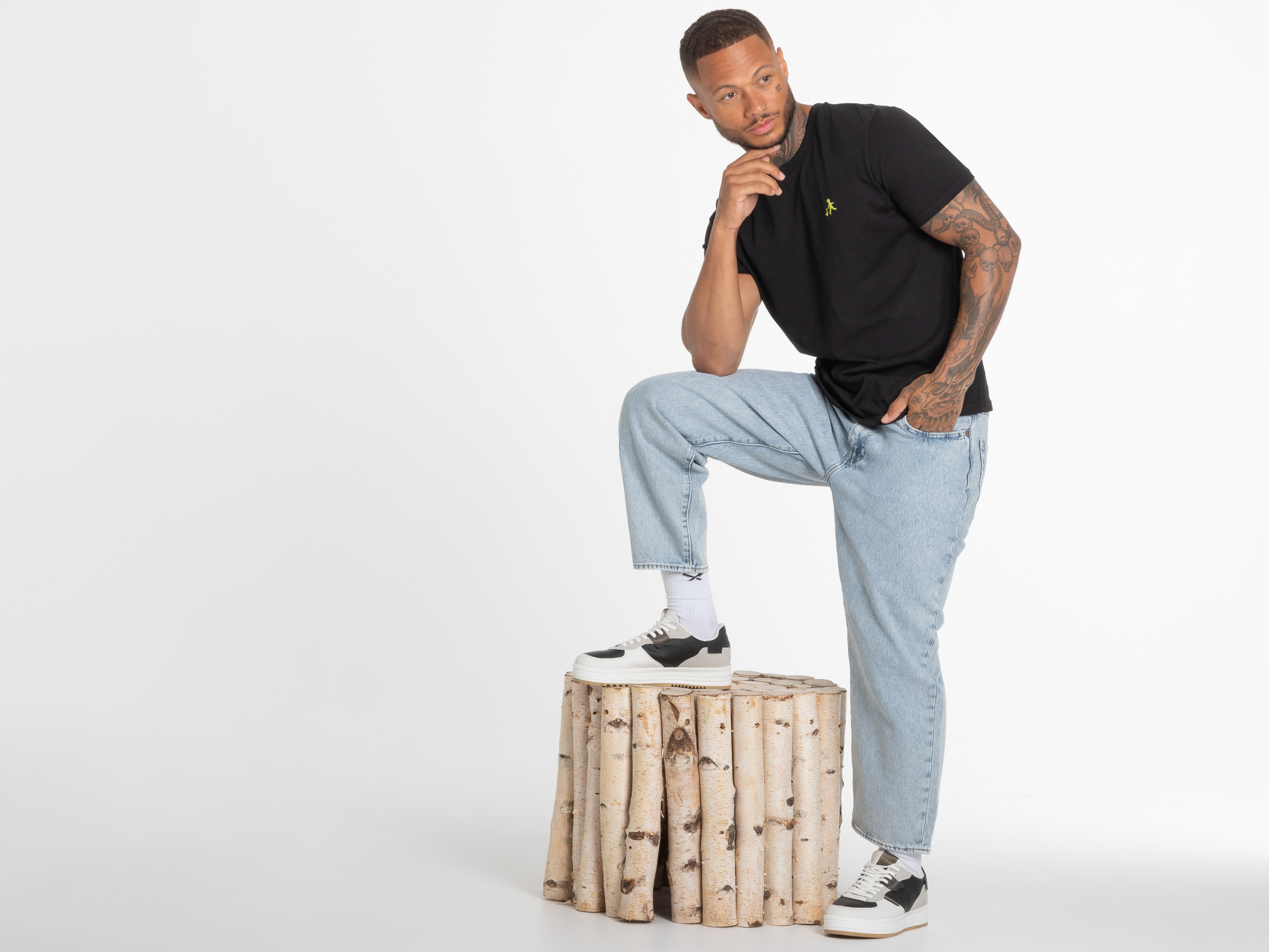 Model steht mit einem Bein auf Birkenhocker, trägt schwarzes Shirt mit Logo – selbstbewusster und dynamischer Auftritt.