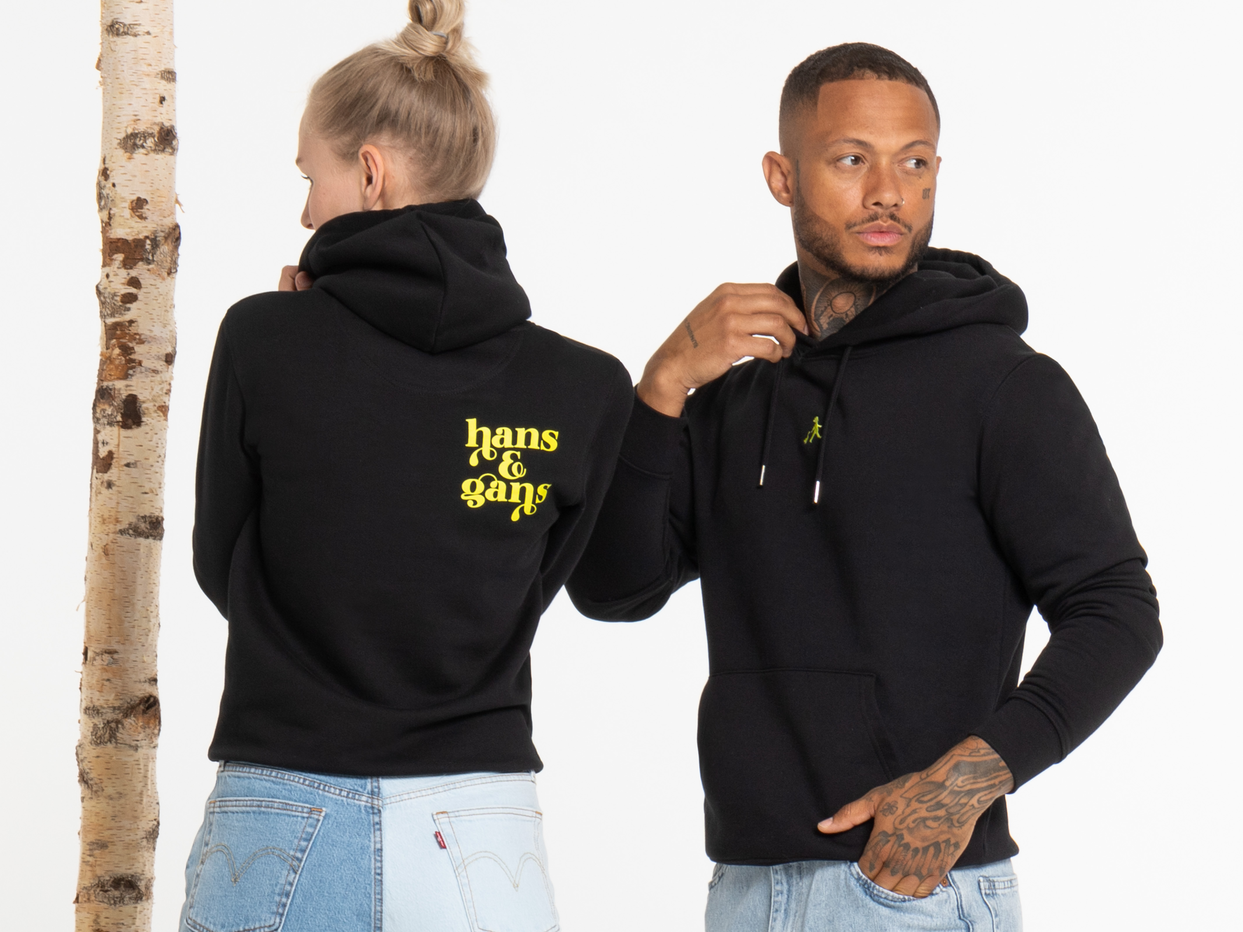Die Frau zeigt den Rückendruck ihres schwarzen Hoodies, der Mann steht seitlich und blickt sie an. Beide lächeln, tragen helle Jeans und schwarze Oberteile.