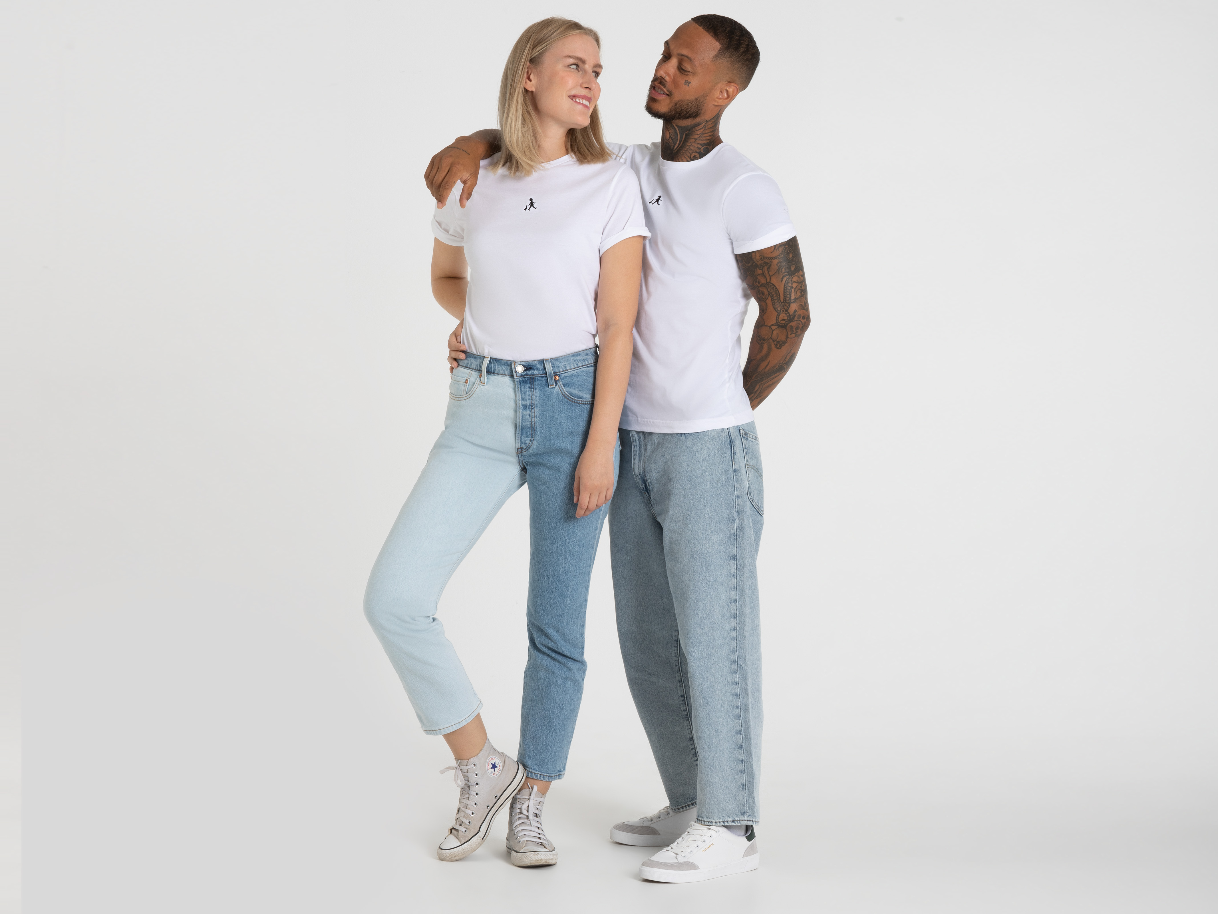 Zwei junge Erwachsene stehen Rücken an Rücken. Beide tragen weiße Oberteile und Jeans. Die Frau hat blondes Haar und lehnt sich leicht zur Seite. Studioaufnahme vor weißem Hintergrund.