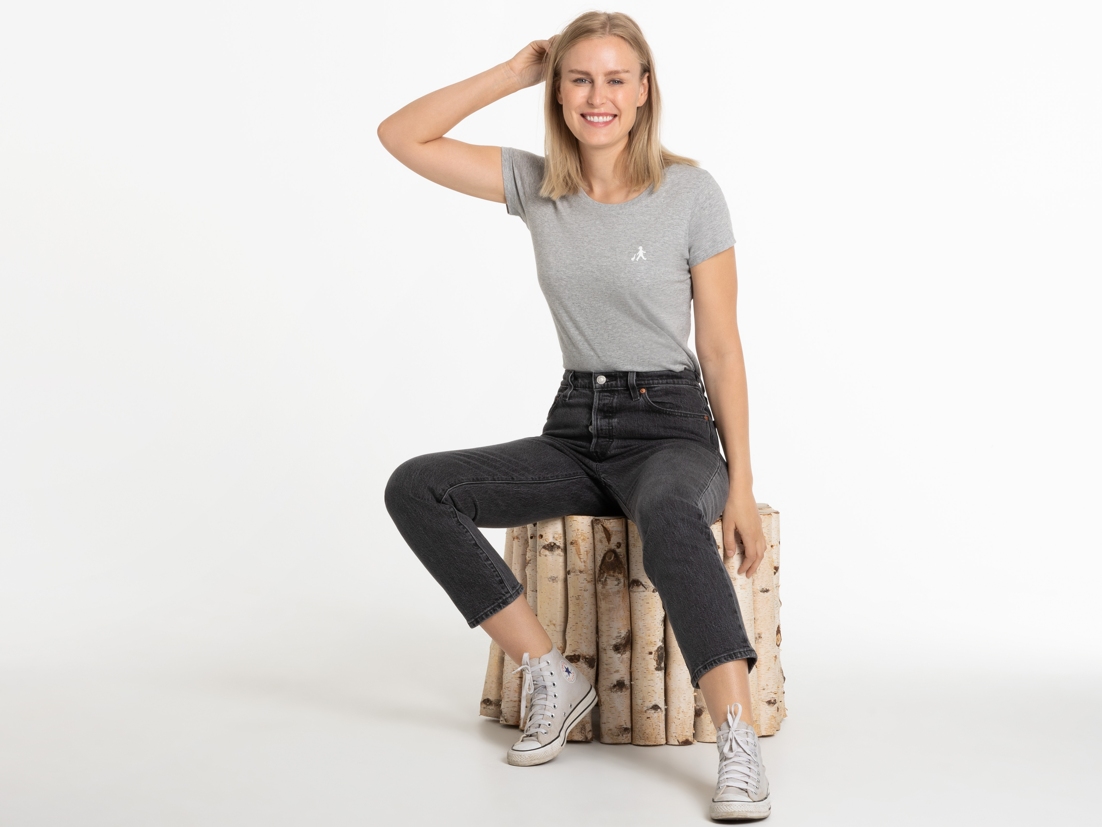 Frau im grauen ‚Hans im Glück‘-T-Shirt, sitzend auf Birkenhocker, Blick zur Seite – entspannt und natürlich.