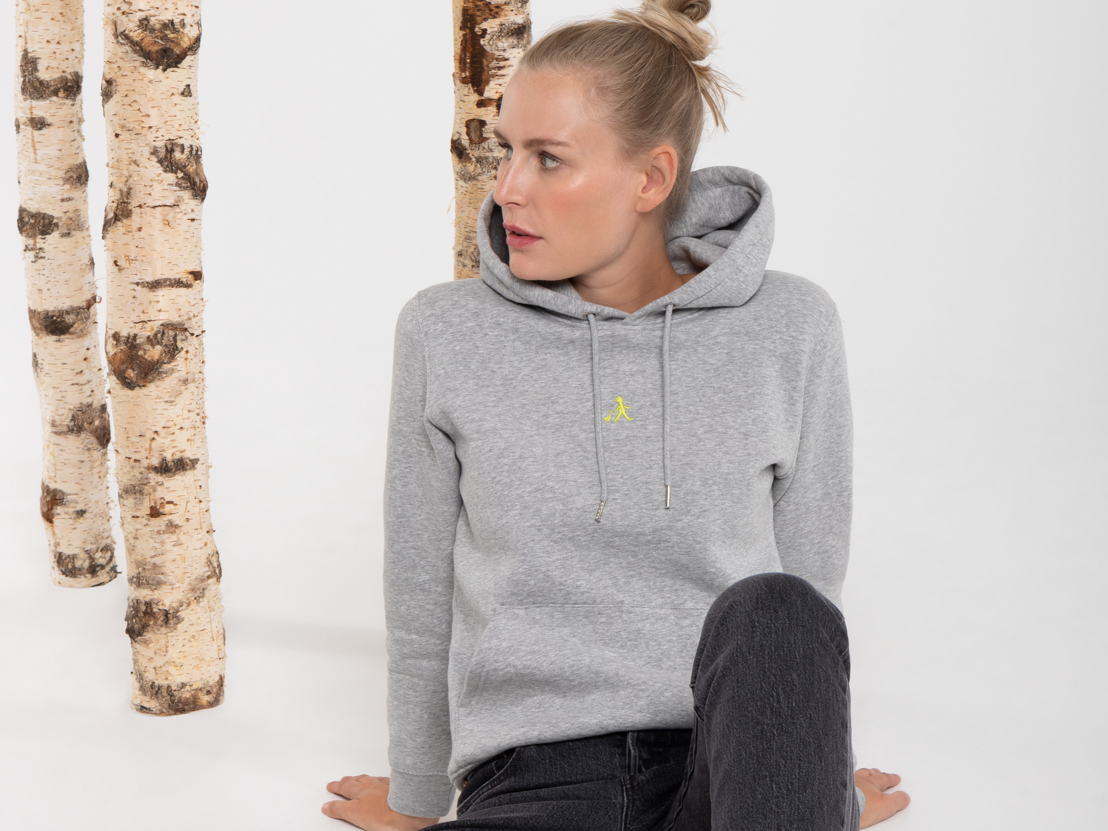 Die Frau sitzt auf dem Boden vor Birkenstämmen, blickt nach links und lächelt leicht. Sie trägt einen grauen Hoodie mit gelbem Logo und eine dunkle Hose.