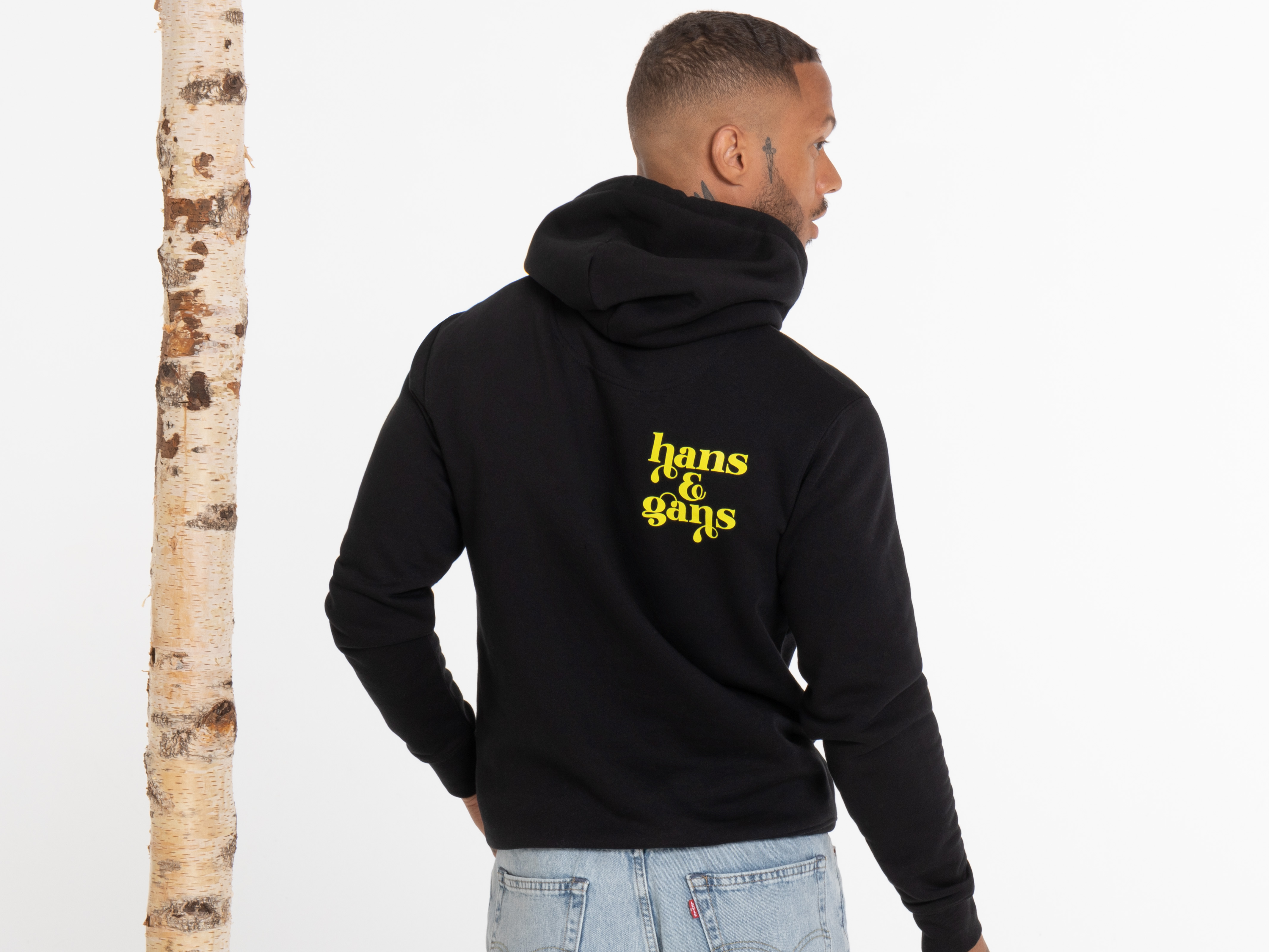Der gleiche Mann ist von hinten im Halbprofil zu sehen, leicht nach rechts gedreht. Auf dem Rücken seines schwarzen Hoodies ist gut sichtbar der gelbe Aufdruck „HANS IM GLÜCK“. Er trägt Jeans, sein rechter Arm hängt locker herab.