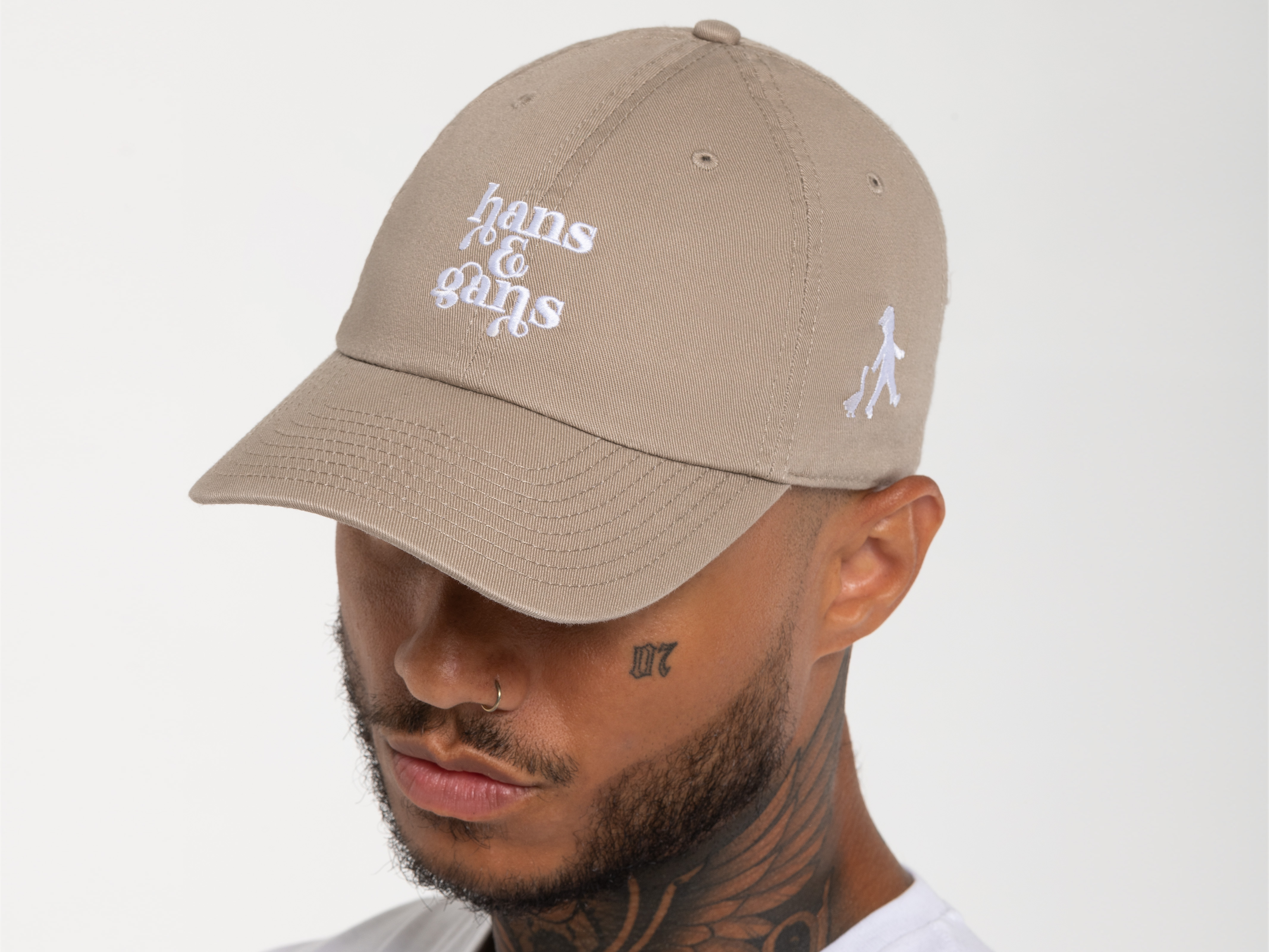 Seitenansicht eines Mannes mit beigefarbener Cap und weißem T-Shirt. Der weiße Schriftzug auf der Cap ist zentral platziert, darunter das Hans-im-Glück-Maskottchen.