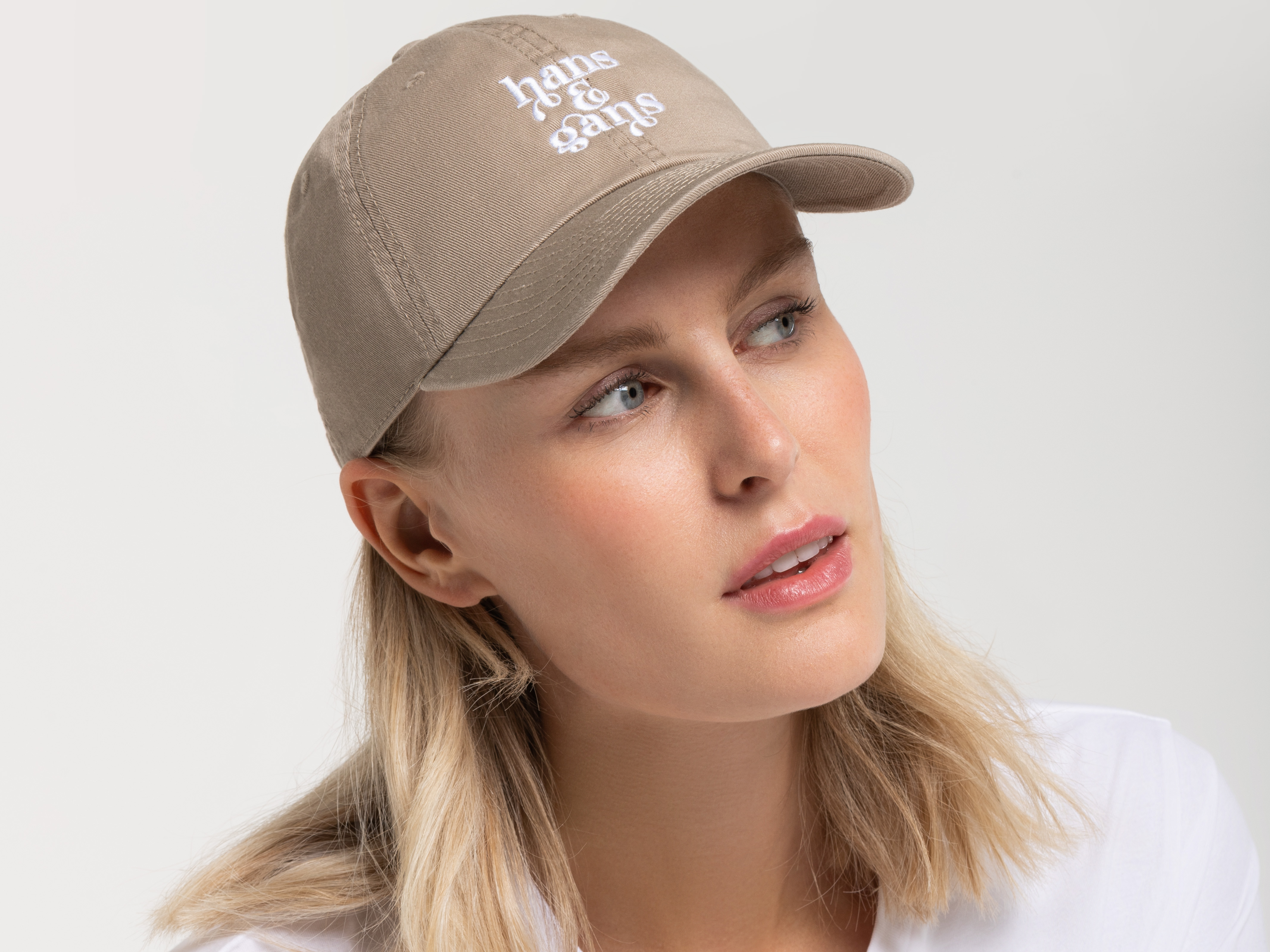 Porträt einer Frau mit hellblonden Haaren, die eine beigefarbene Cap mit weißem Sticklogo trägt. Sie blickt leicht nach oben, trägt ein weißes T-Shirt.