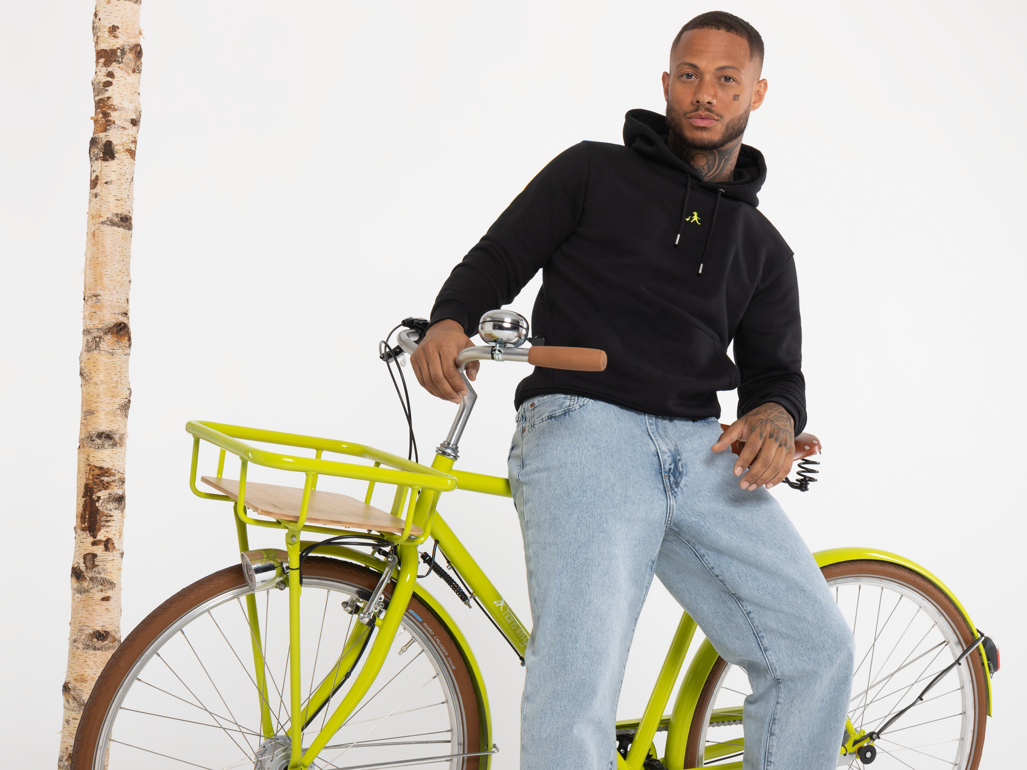 Ein Mann steht seitlich hinter einem gelben Fahrrad mit Holzgepäckträger. Er trägt einen schwarzen Hoodie und Jeans, sein Blick geht aus dem Bild.
