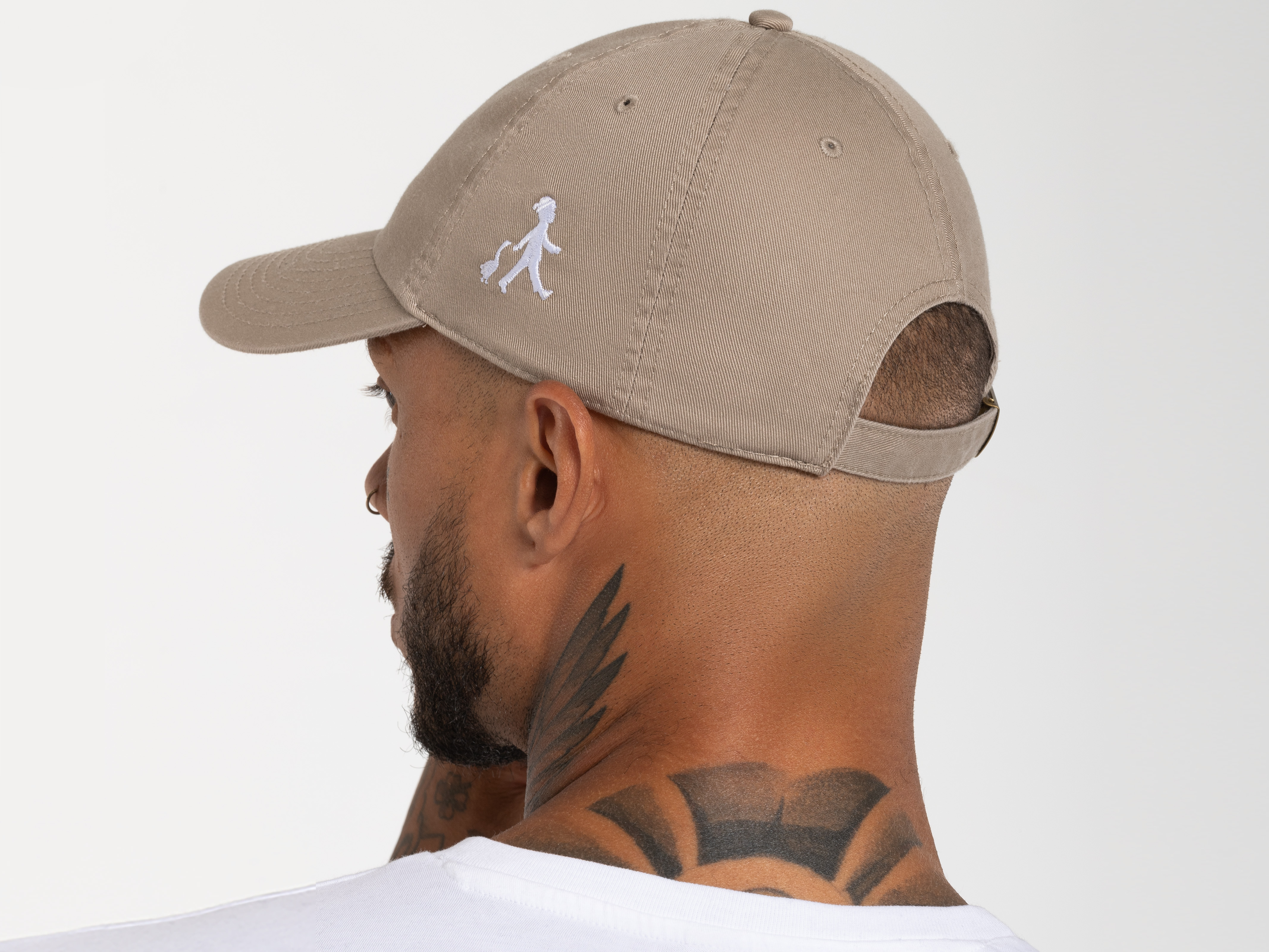 Rückansicht der beigefarbenen Cap, getragen von einem Mann mit kurzem Haar und Nackentattoo. Der verstellbare Riemen und das Logo auf der Seite sind erkennbar.