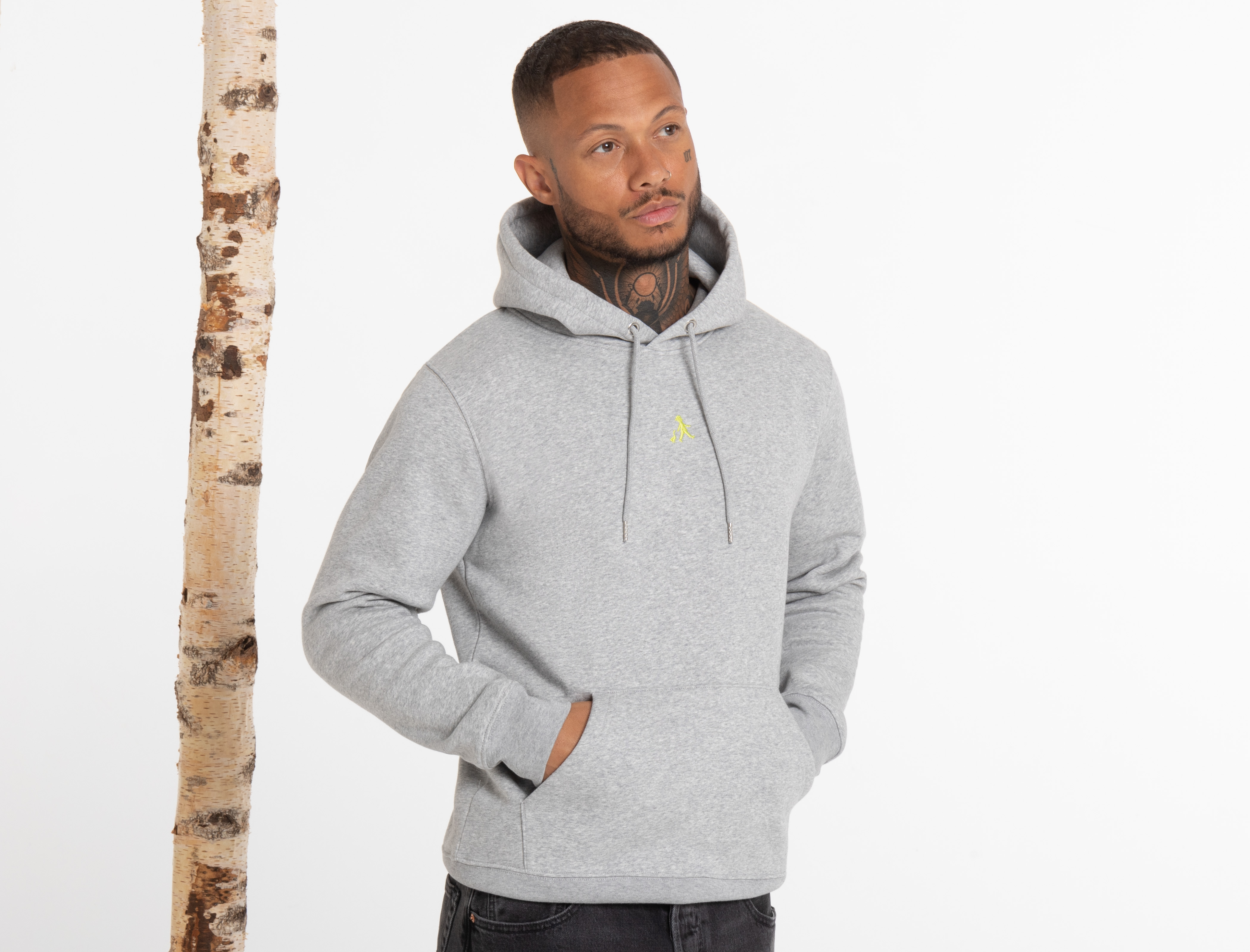 Der Mann blickt leicht nach links, trägt einen grauen Hoodie mit Kängurutasche und eine schwarze Hose. Zwei Birkenstämme stehen hinter ihm.