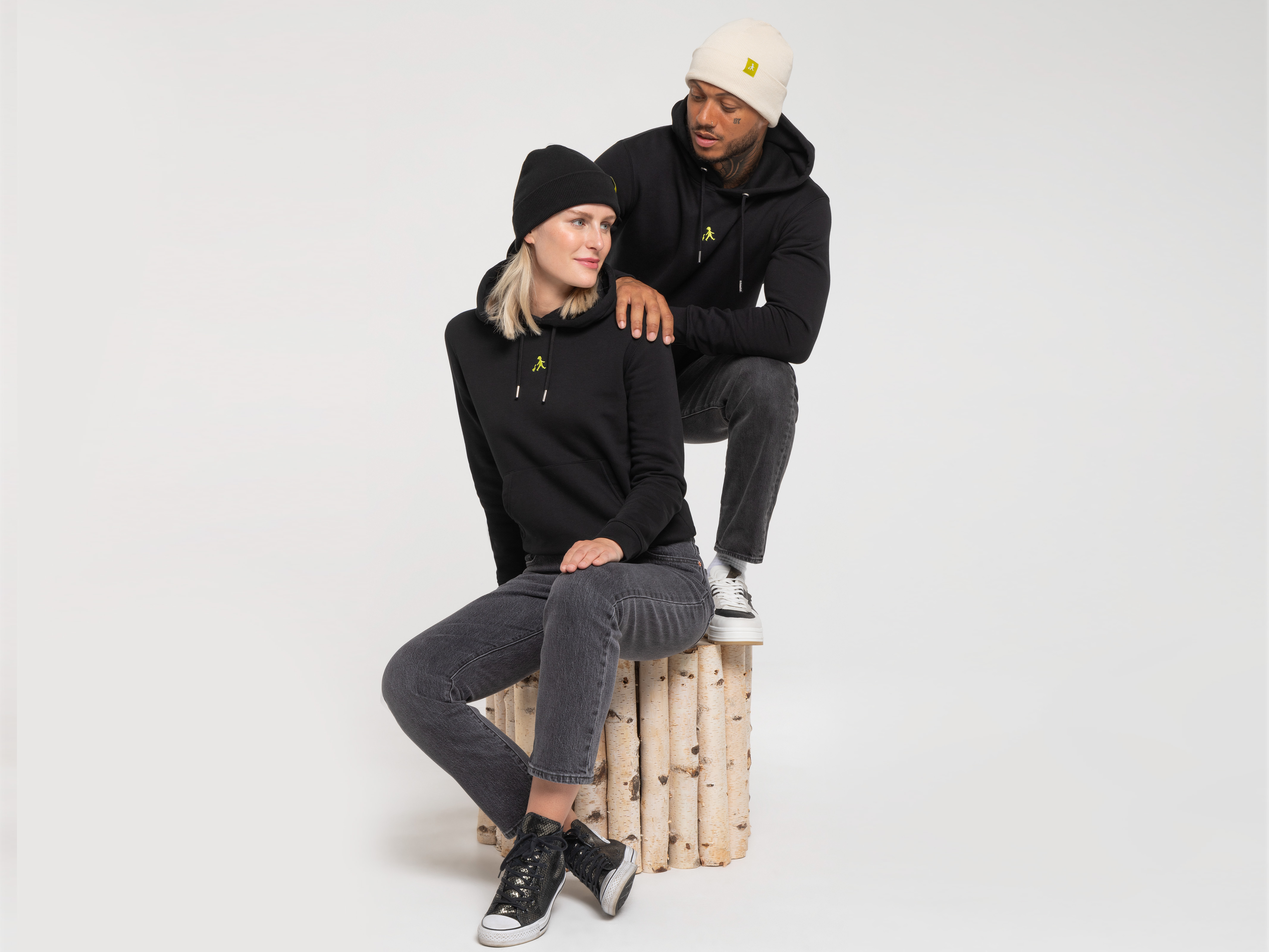 Zwei Personen sitzen auf einem Holzhocker, beide in schwarzem Hoodie. Die Frau trägt eine beigefarbene Beanie, der Mann schaut ruhig zur Seite.