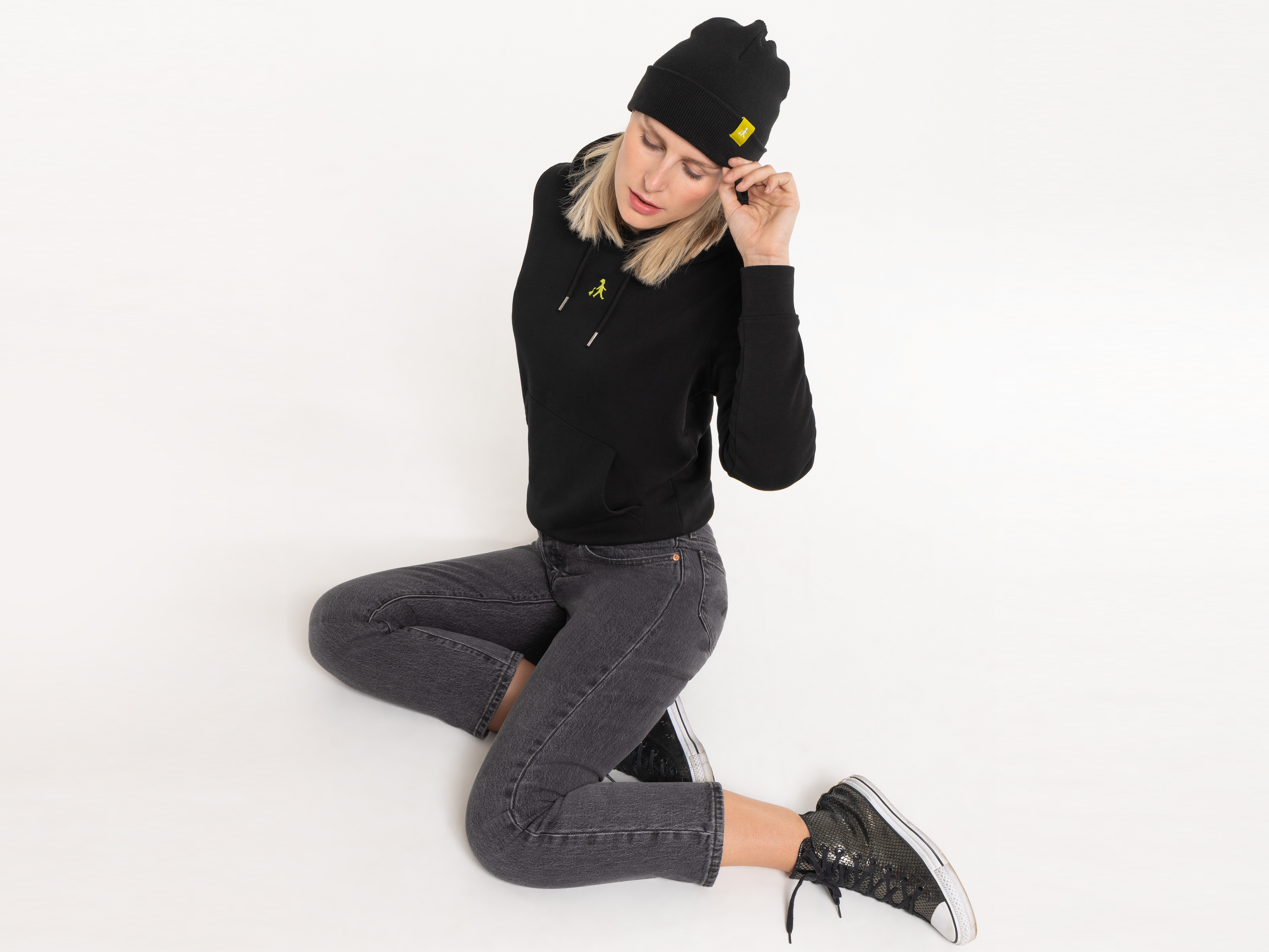 Die Frau kniet auf dem Boden, trägt eine schwarze Beanie und schwarzen Hoodie. Ihre Pose wirkt verspielt und modisch.