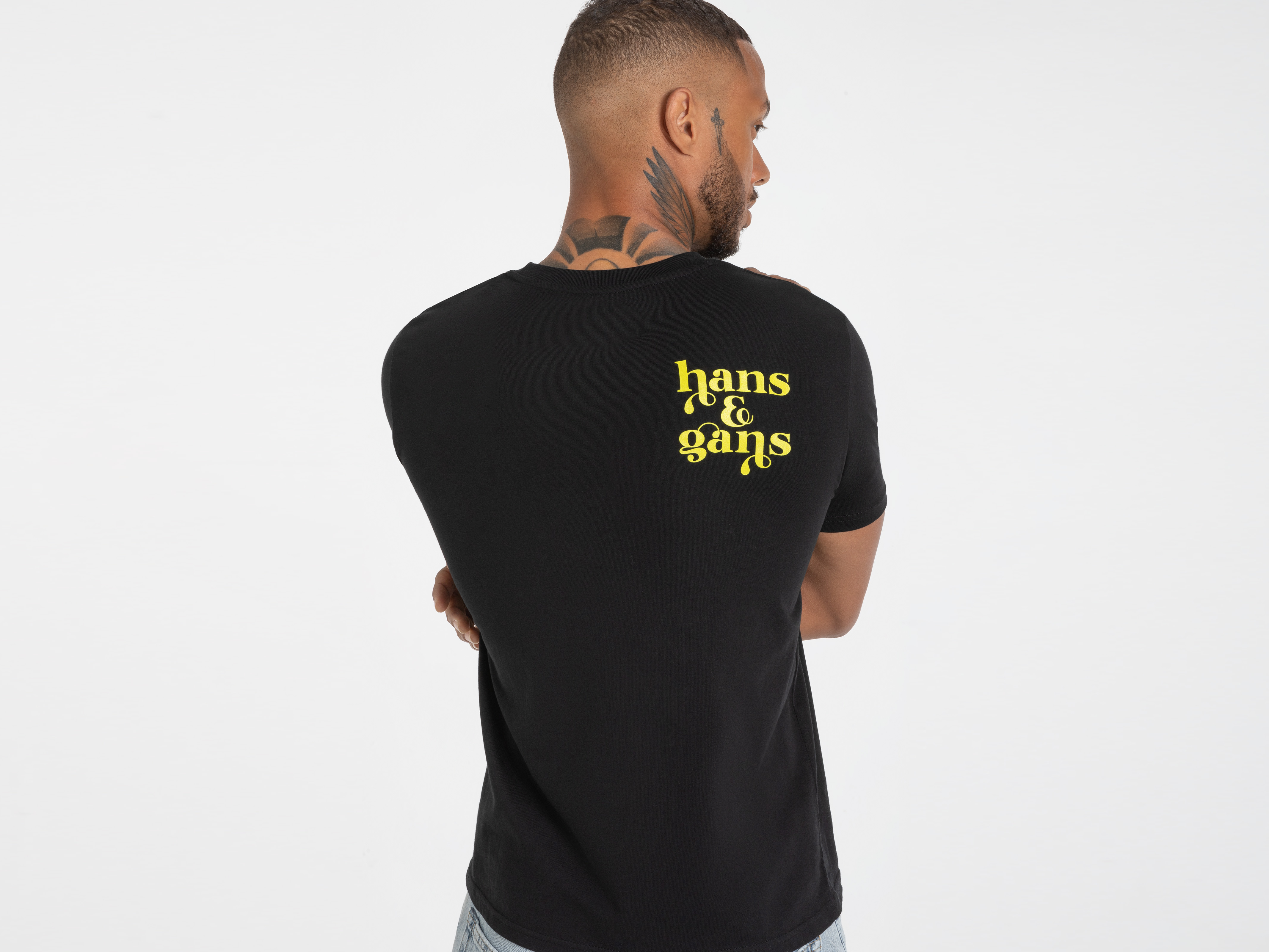 Schwarzes T-Shirt von hinten mit markantem gelbem Rückendruck ‚Hans im Glück‘ – authentischer Streetstyle.