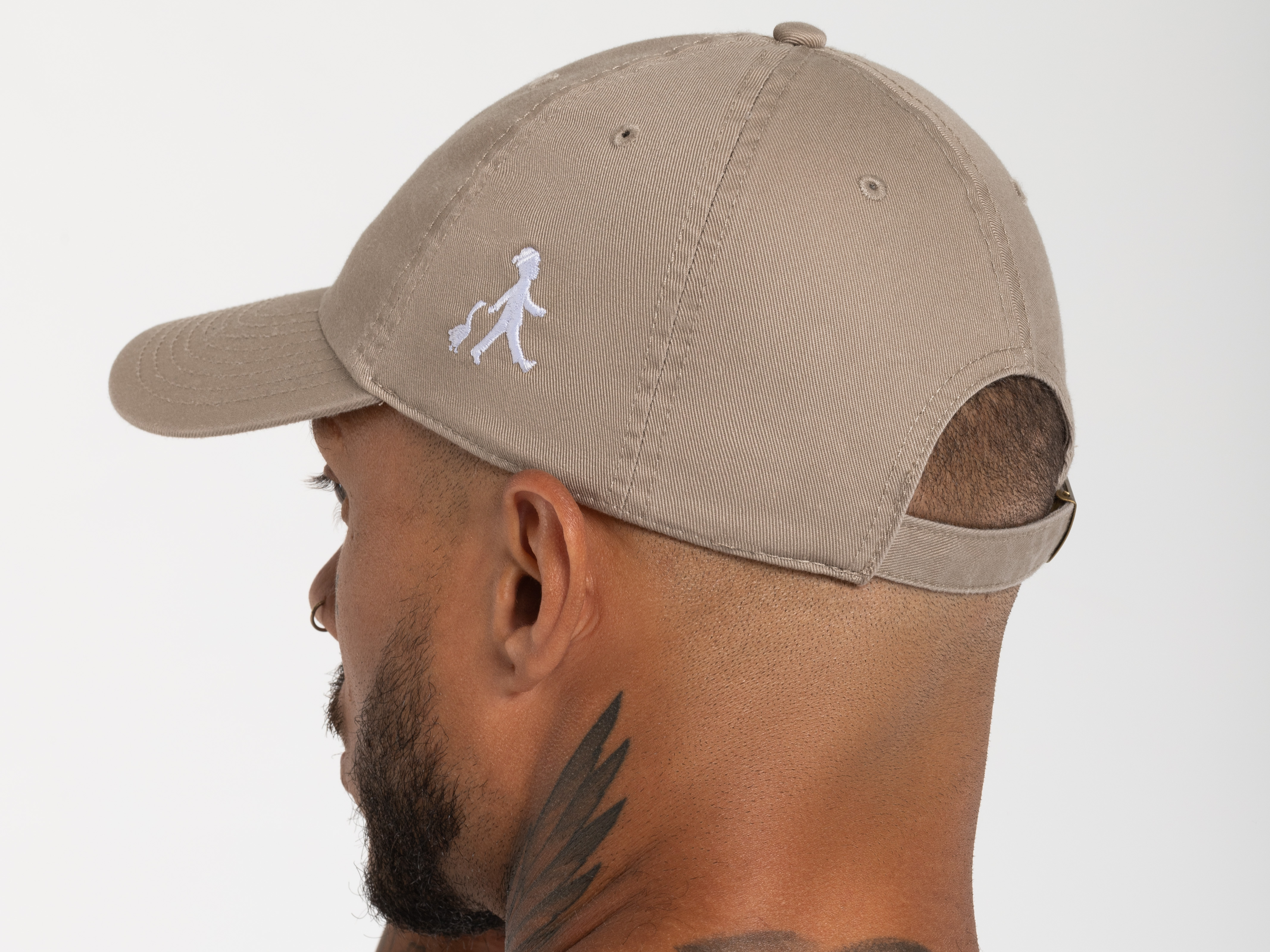 Seitliche Nahaufnahme eines Mannes mit beigefarbener Cap, das gestickte Maskottchen-Logo ist deutlich sichtbar. Er trägt ein schwarzes Oberteil.