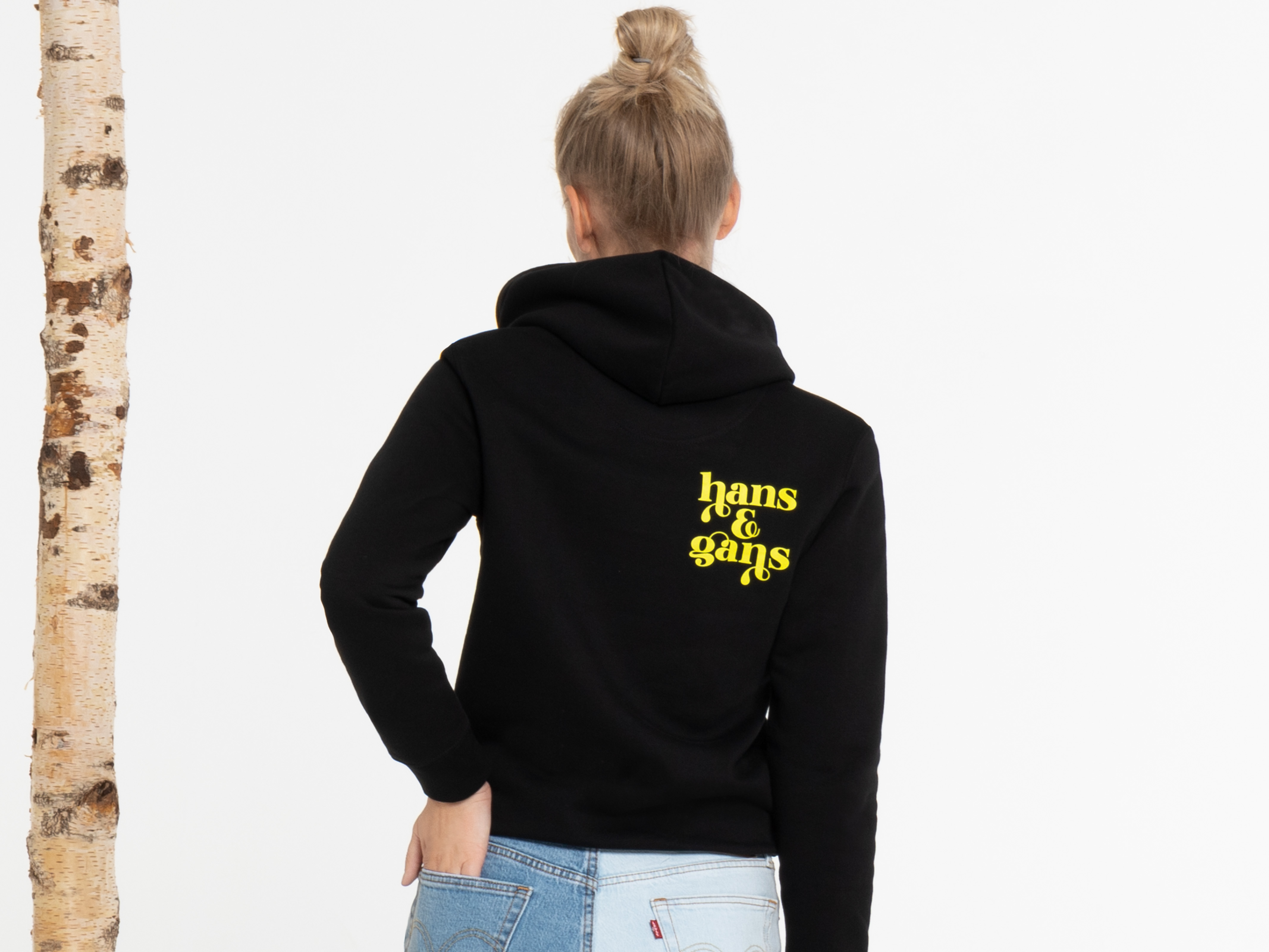 Rückenansicht einer Frau mit schwarzem Hoodie. Der gelbe Aufdruck ist zentral zu sehen, die Haare sind zum Dutt gebunden. Studioaufnahme mit Holz-Elementen.