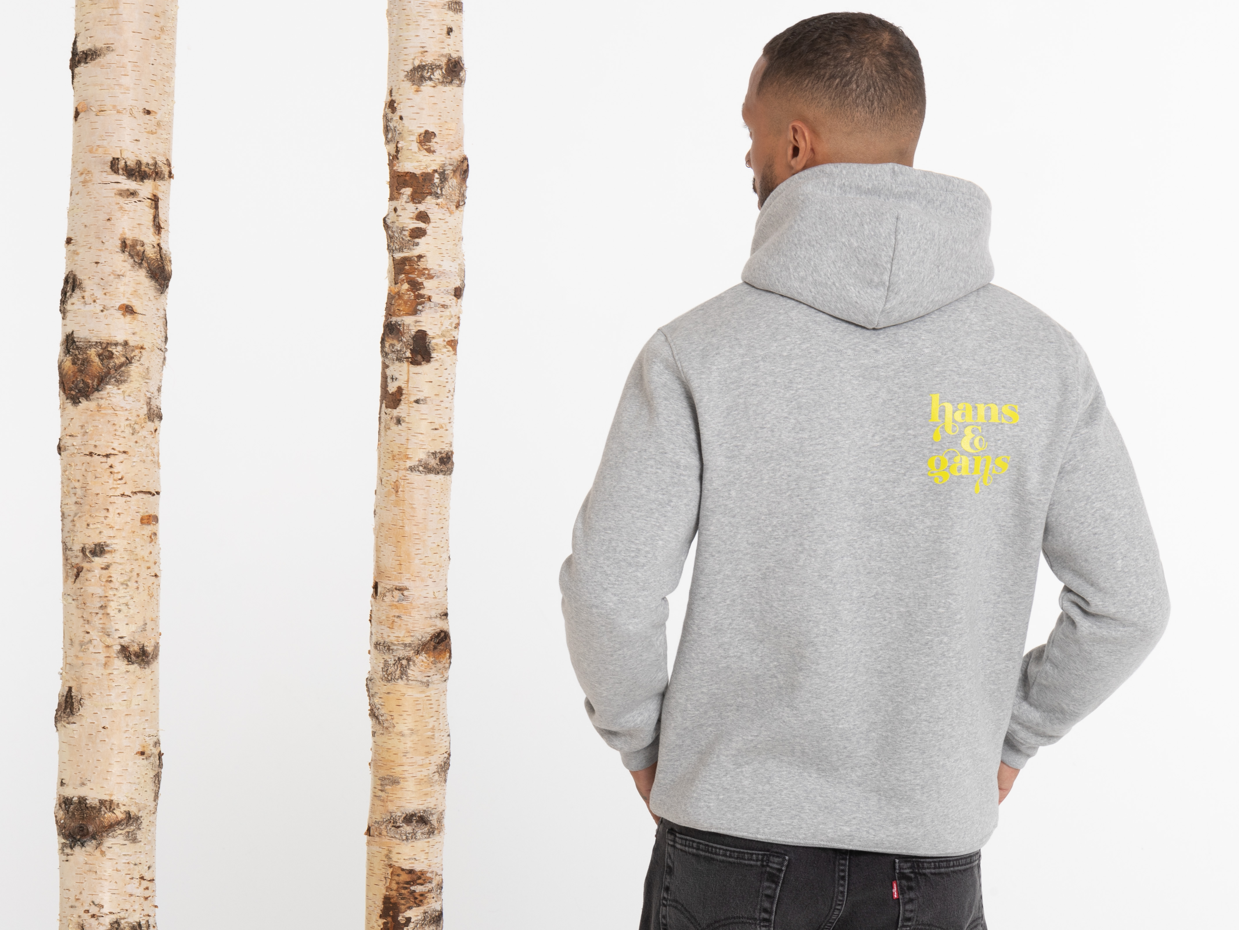 Rückansicht des Mannes in grauem Hoodie mit großem gelbem Print auf dem Rücken. Die Arme hängen locker, er steht zwischen zwei Birkenstämmen.