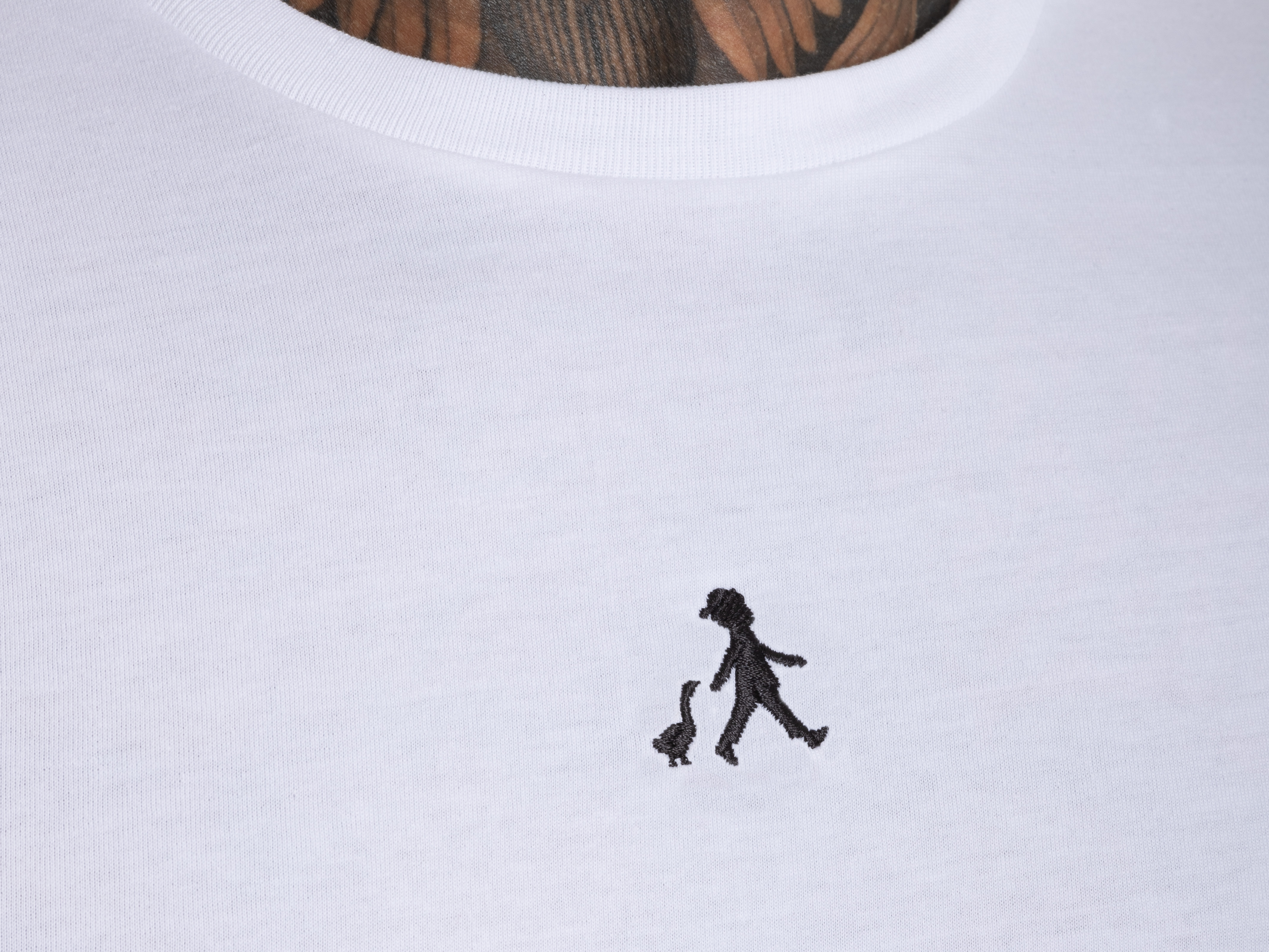 Detailaufnahme des ‚Hans im Glück‘-Logos auf T-Shirt mit sichtbarem Tattoo im Hintergrund – Kombination aus Design und Persönlichkeit.