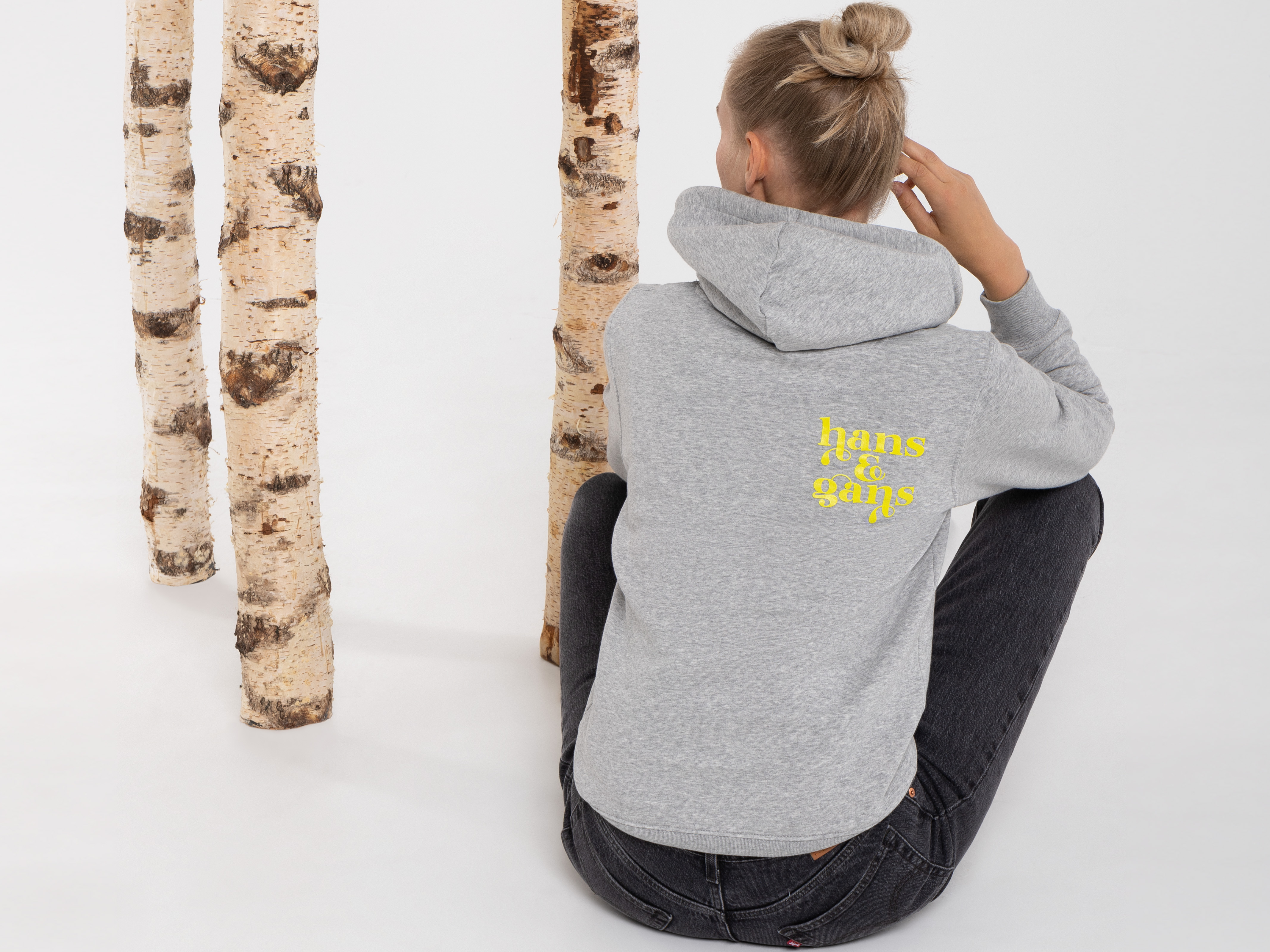 Rückansicht der Frau im grauen Hoodie mit großem gelbem Rückenprint. Sie sitzt am Boden mit überkreuzten Beinen, drei Birkenstämme im Hintergrund.