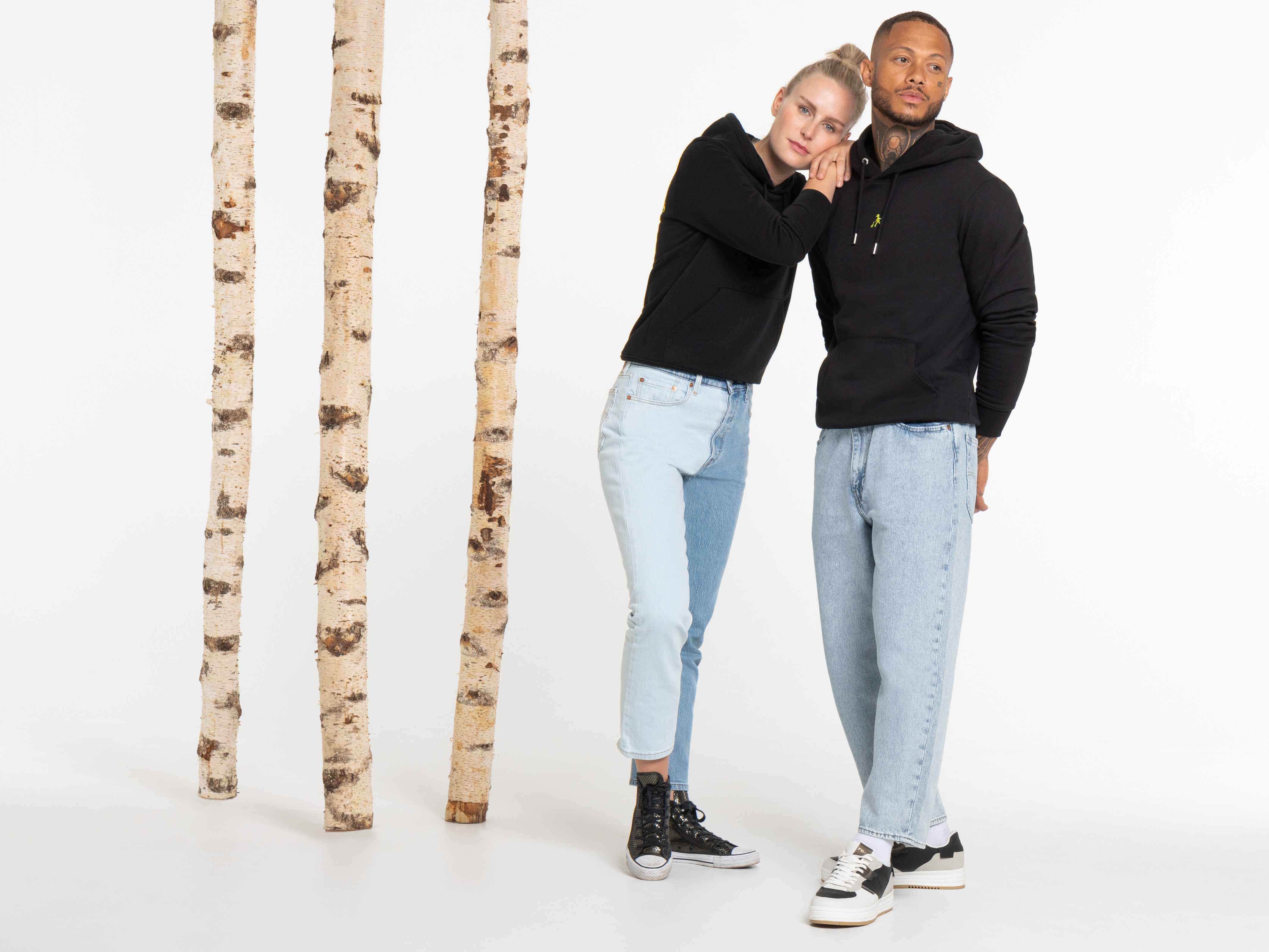 Zwei Personen stehen seitlich nebeneinander, tragen schwarze Hoodies und helle Jeans. Eine legt den Arm um die Schulter der anderen. Beide lächeln leicht.